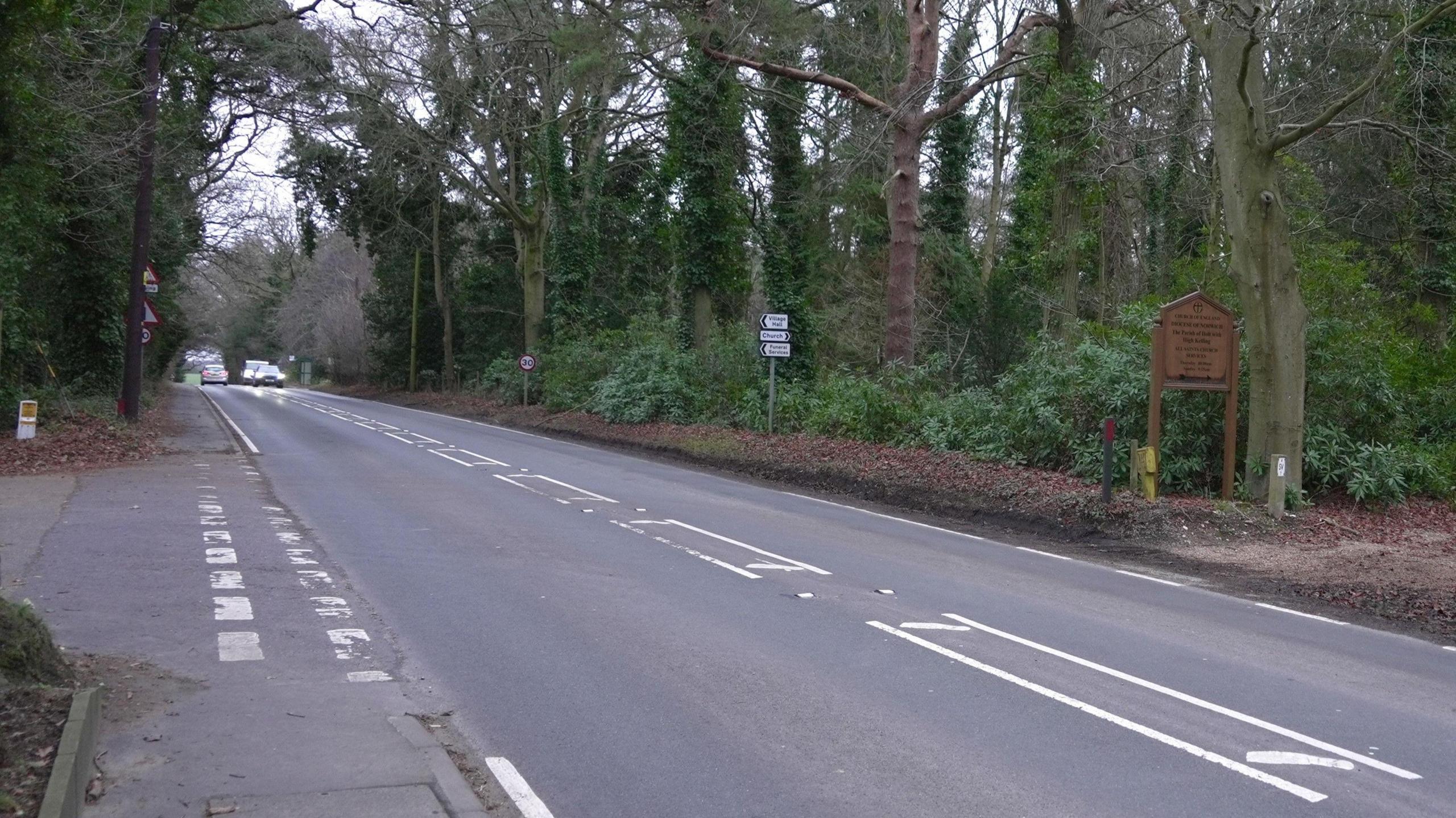 Another view of Avenue Road in High Kelling. Vehicles can be seen driving in both directions further up the road with various signs next to the road. Trees line either side of the road. 