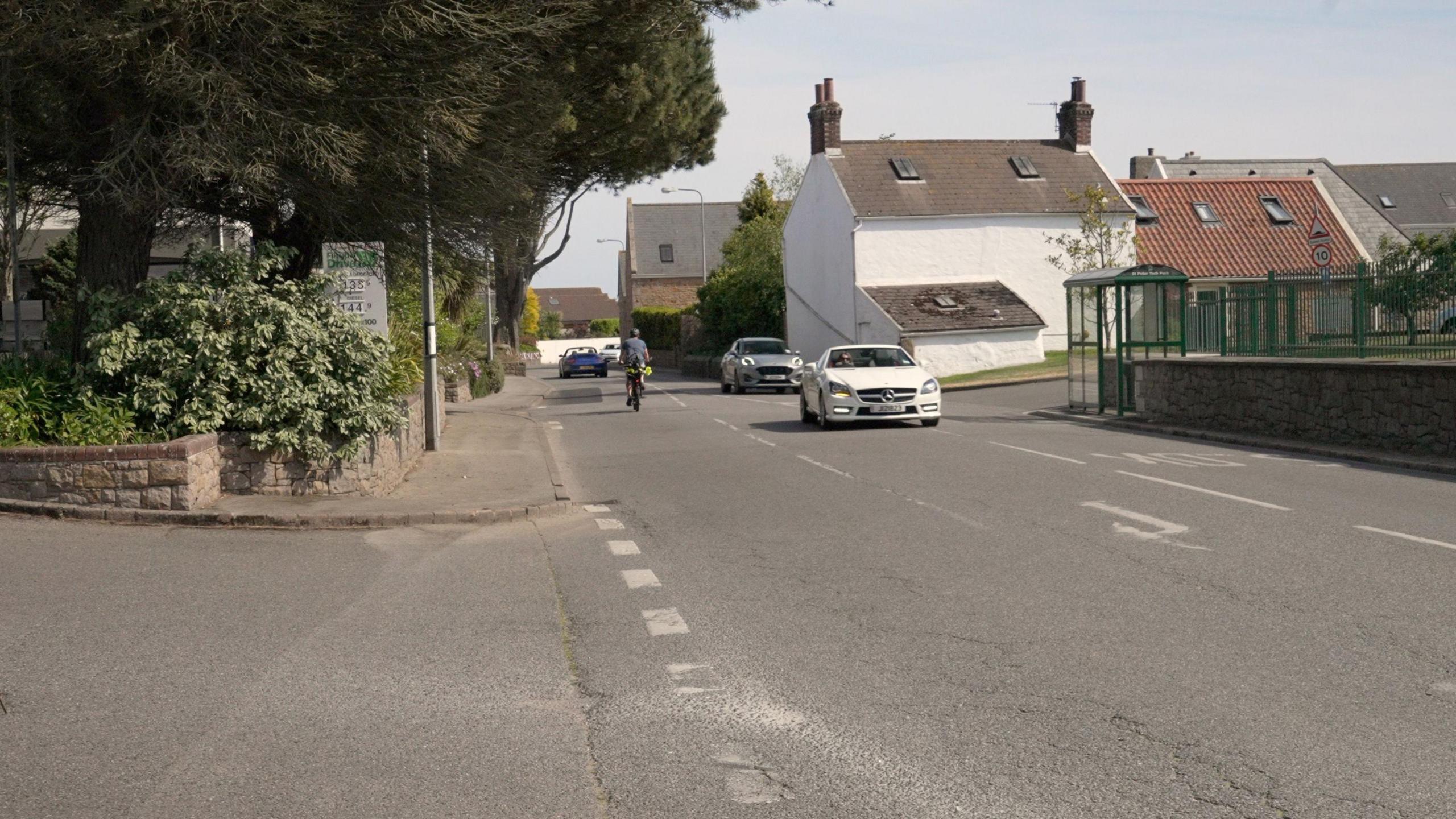 A road in St Peter, with a petrol station on the left hand side of the road - largely covered in trees, and houses and a bus station on the right hand side. There is a right-hand turn lane in the middle of the road. A white car is indicating into the turn lane, with a silver car travelling behind it. A cyclist and blue car are on the left hand side of the road travelling away from the camera.