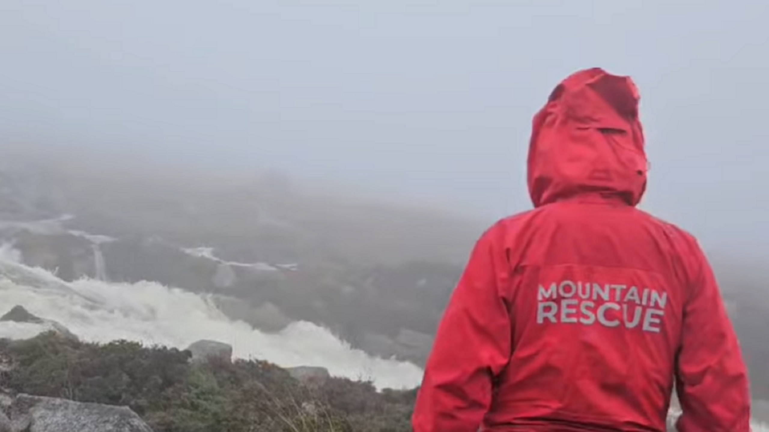 A person is dressed in a bright read jacket with its hood pulled up and the words Mountain Rescue printed on the back. The person is looking towards a swollen river in a misty mountain landscape.