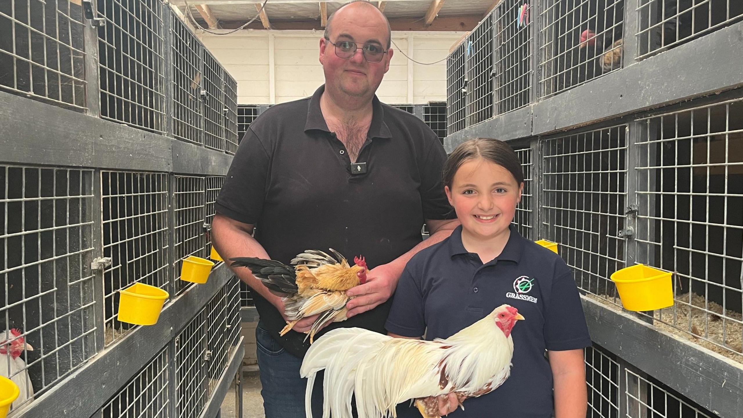 Alannah (right) stands with her dad Neal (left).Neal is wearing a black polo shirt and jeans and glasses. Alannah is wearing a navy polo shirt with a white and green logo. Neal is holding a small brown and black chicken and Alannah is holding a larger white chicken with long tail feathers. They are both smiling at the camera. They are standing inside an indoor chicken coop with multiple enclosures. There are metal grated gages with bright yellow feed boxes on the front.