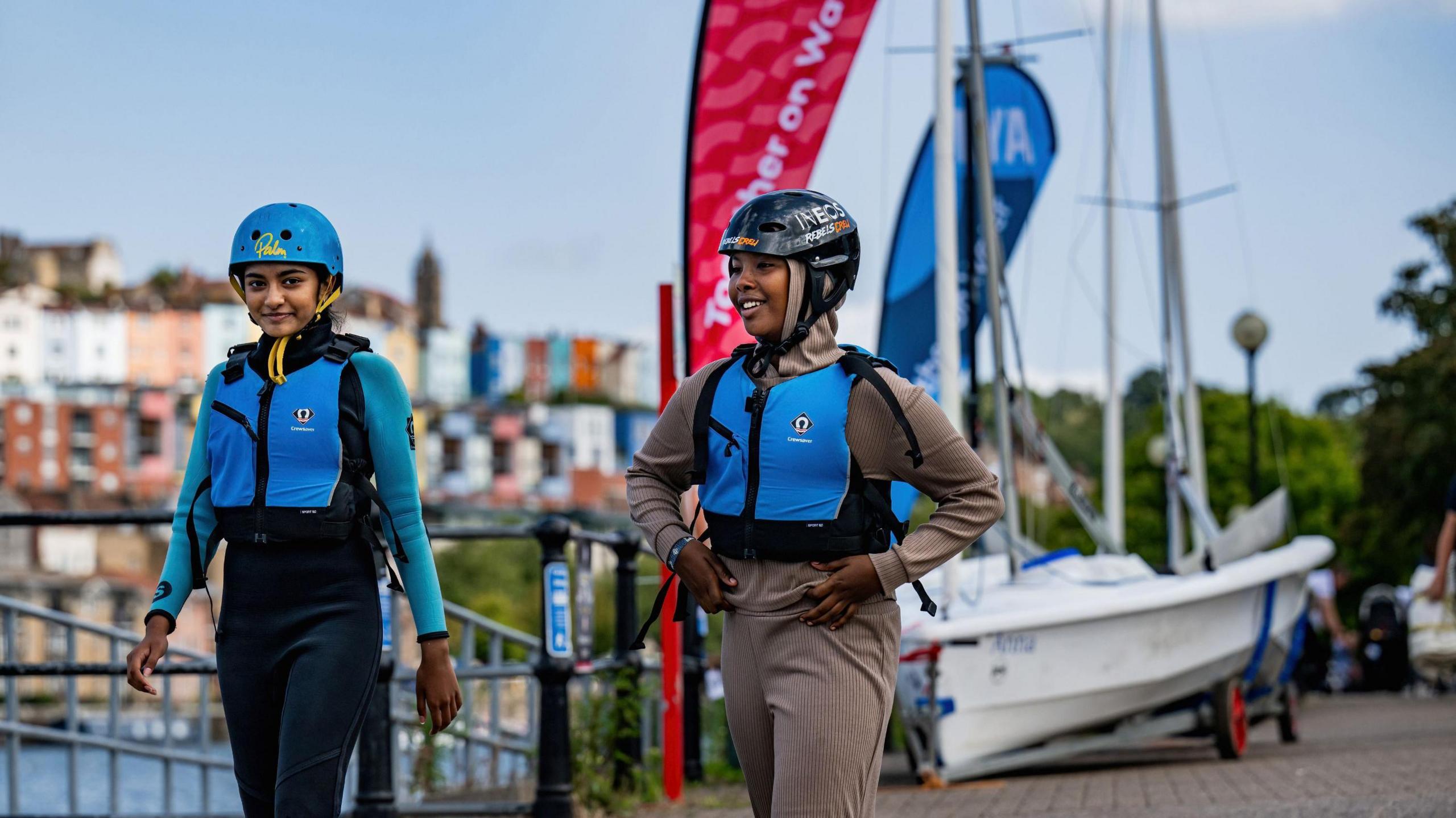 Two girls walking along the the harbour with a small sailing boat behind them. They're wearing blue lifejackets and helmets