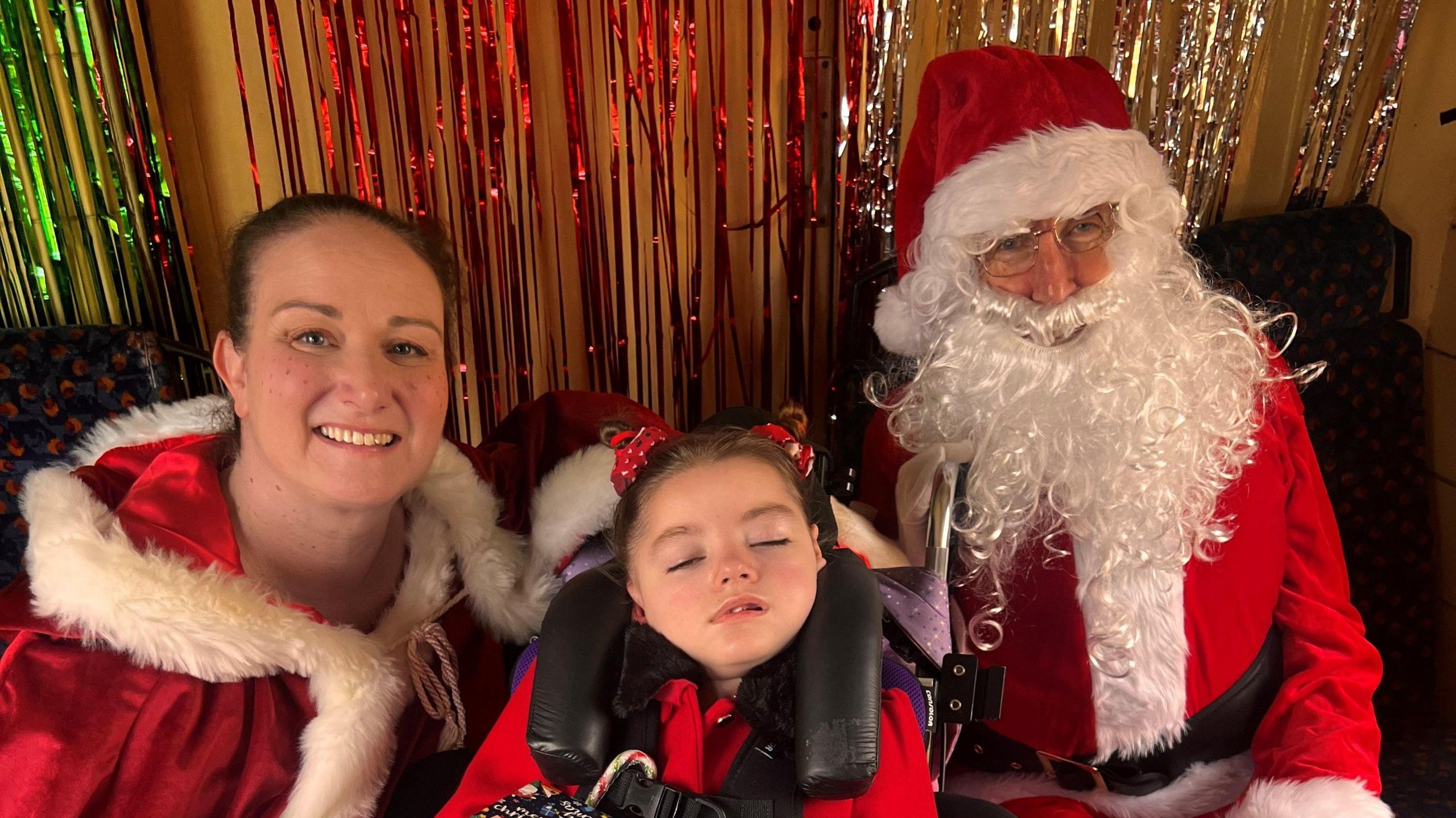 A girl is in the middle with her eyes cut, a woman in a red top is on the left smiling at the camera and a man with glasses was dressed as Santa on the right.