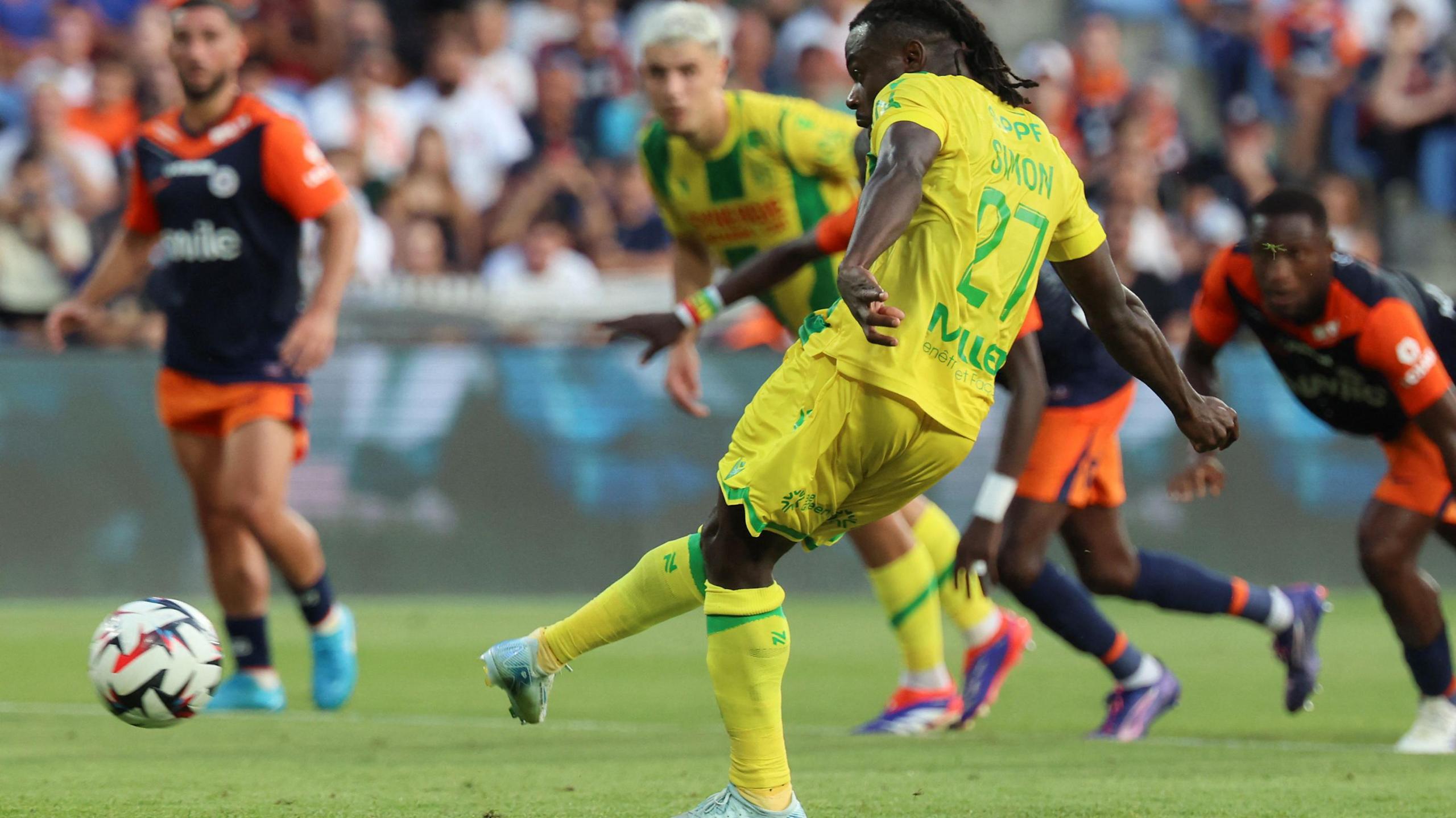 Moses Simon, with most of his body turned away from the camera, sends a side-footed penalty towards the left of the picture and the goal