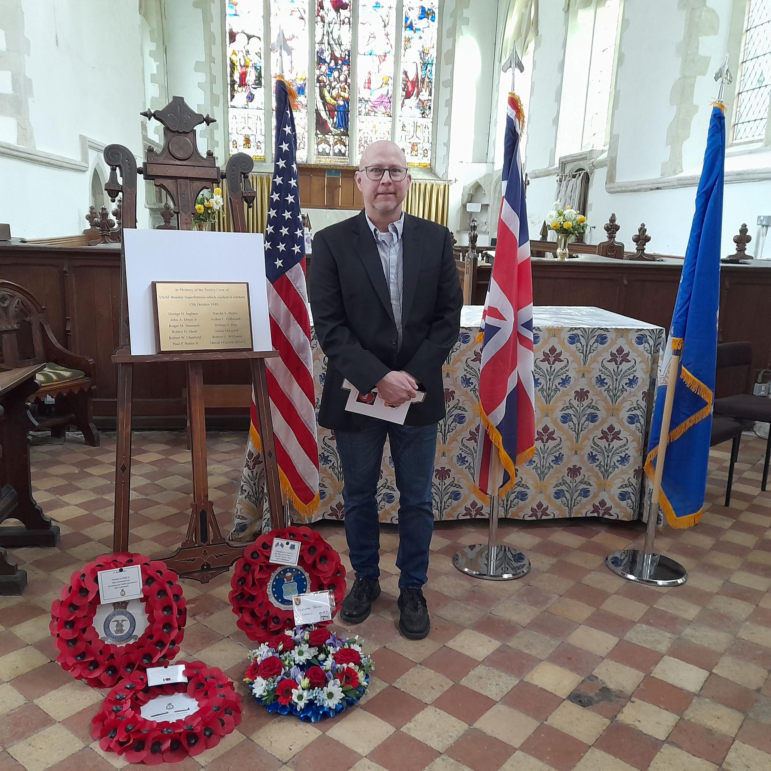 A man with glasses and a dark jacket stands in a church with the flags of USA, GB and a military standard. He holds an order of service and there are floral and poppy wreaths at his feet. A plaque commemorating the crash is on a stand next to him.