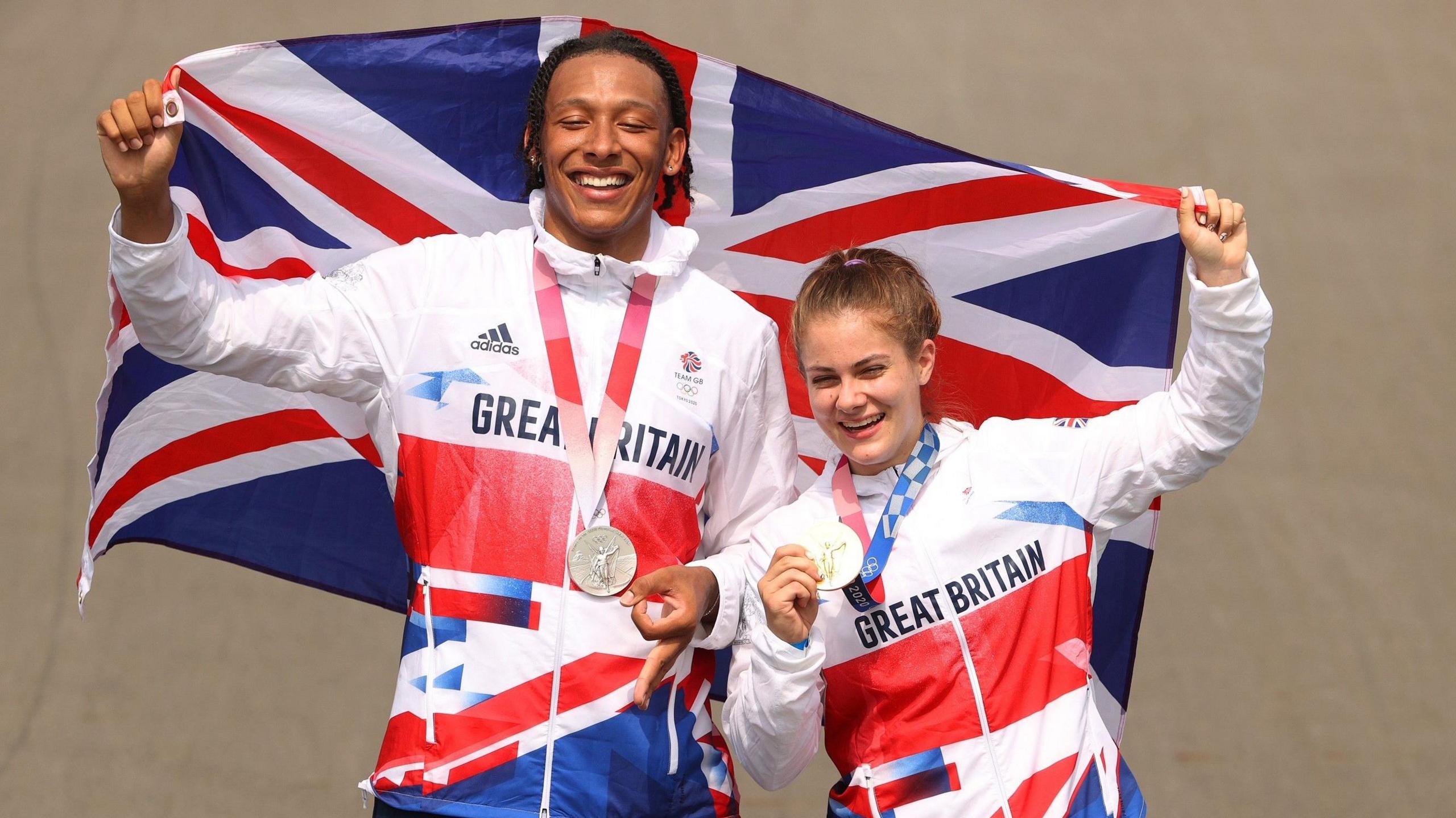 Picture of Britain's BMX Racing medallists from Tokyo 2020 Olympic Games. Kye White on the left with his silver medal and Beth Shriever on the right with her gold medal - both smiling while carrying a Union flag