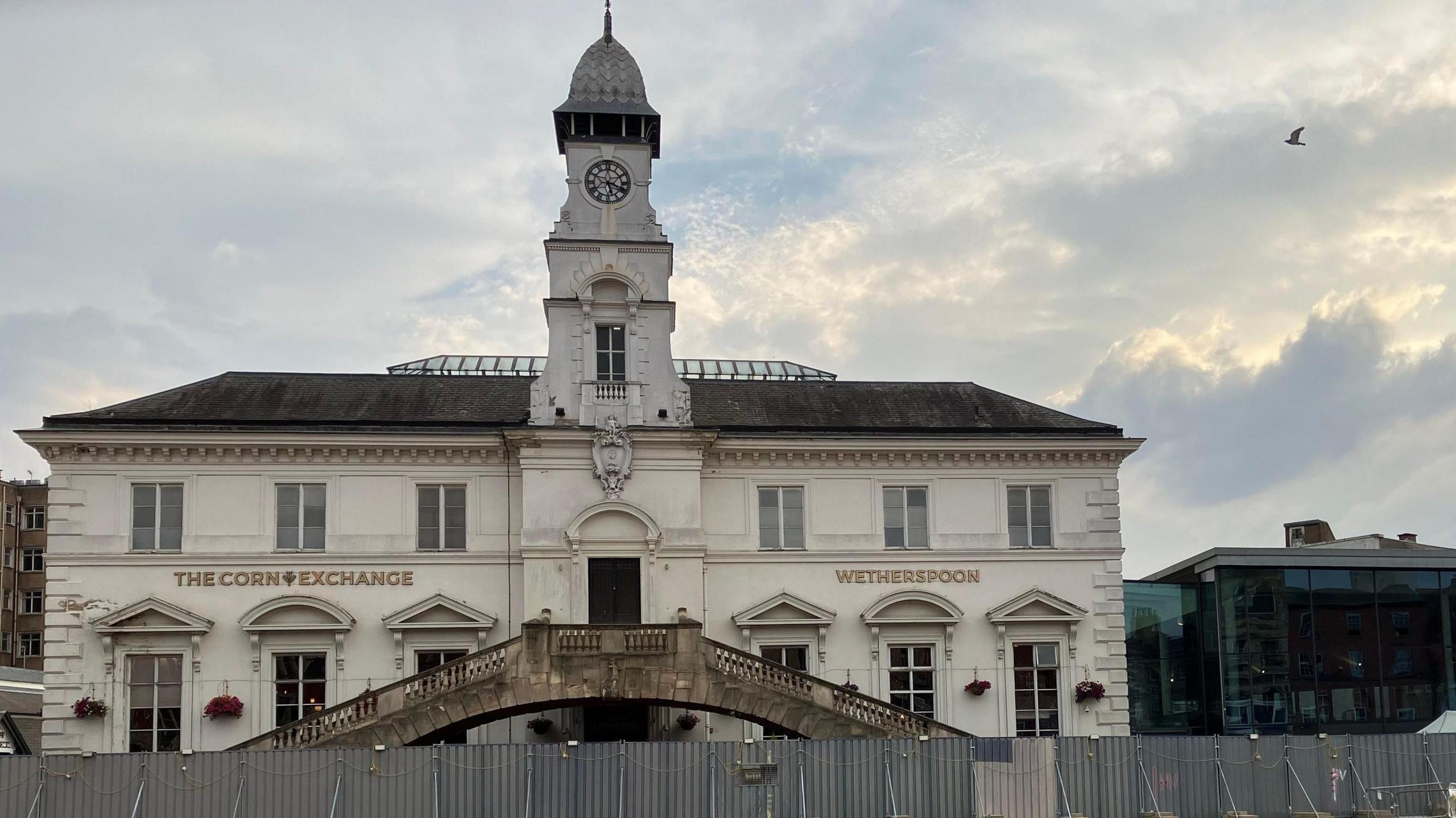 The Rialto Bridge, made from stone with two sets of staircases that meet in the middle, in front of the white Corn Exchange building. The building has a clock tower in the middle. There is temporary metal fencing in front of the staircase.