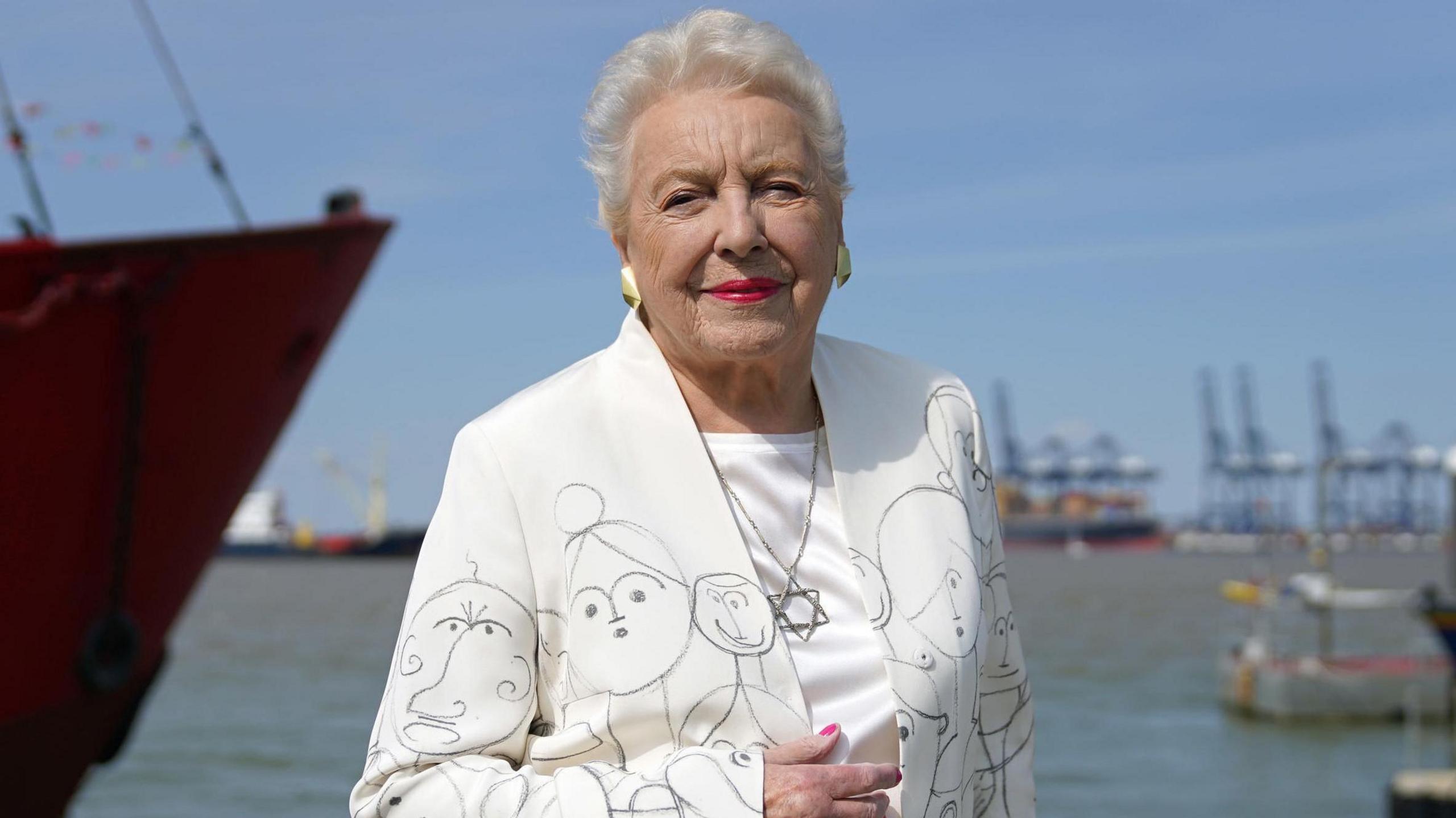 Dame Stephanie Shirley poses infront of the sea in a white suit covered in drawings of children and a Star of David necklace at the unveiling of a memorial to Kindertransport on the quayside at Harwich in 2022