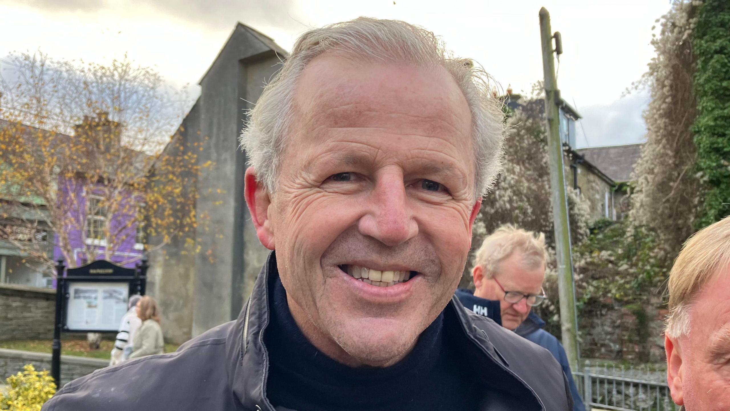 Sean Fitzpatrick who has grey hair combed back from his forehead smiles to the camera. He is wearing a back jumper and black jacket