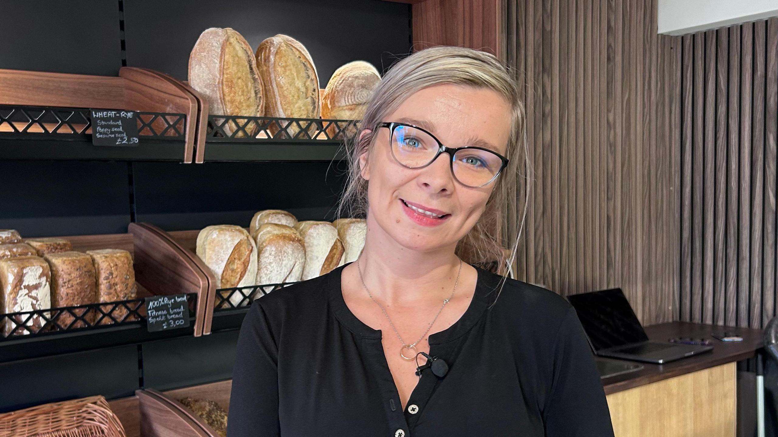 Kate Oslizlok stands in Eunice Artisan Bakery in Kettering. She is in a black top and is wearing spectacles. Behind her are various loaves of bread and a counter with a laptop standing on it.