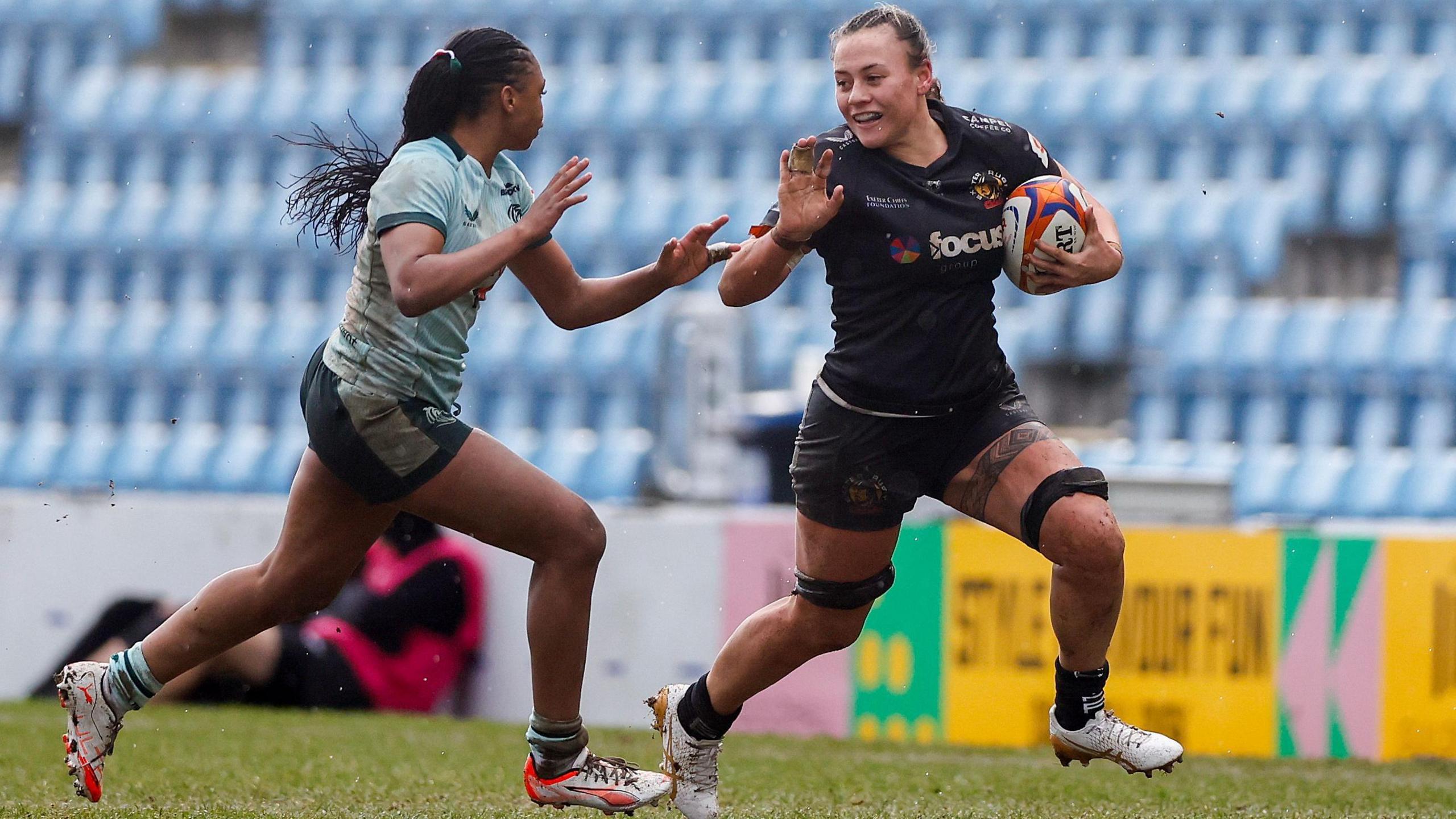 Exeter Chiefs Women rugby football union player Maddie Feaunati holds the ball while holding past a Leicester Tigers Women player during a Women's Premiership Rugby match on grass. Feaunati is wearing a predominantly black kit with white lettering, while her opponent is wearing a light green shirt with dark green trim and dark green shorts. Both players are wearing white boots