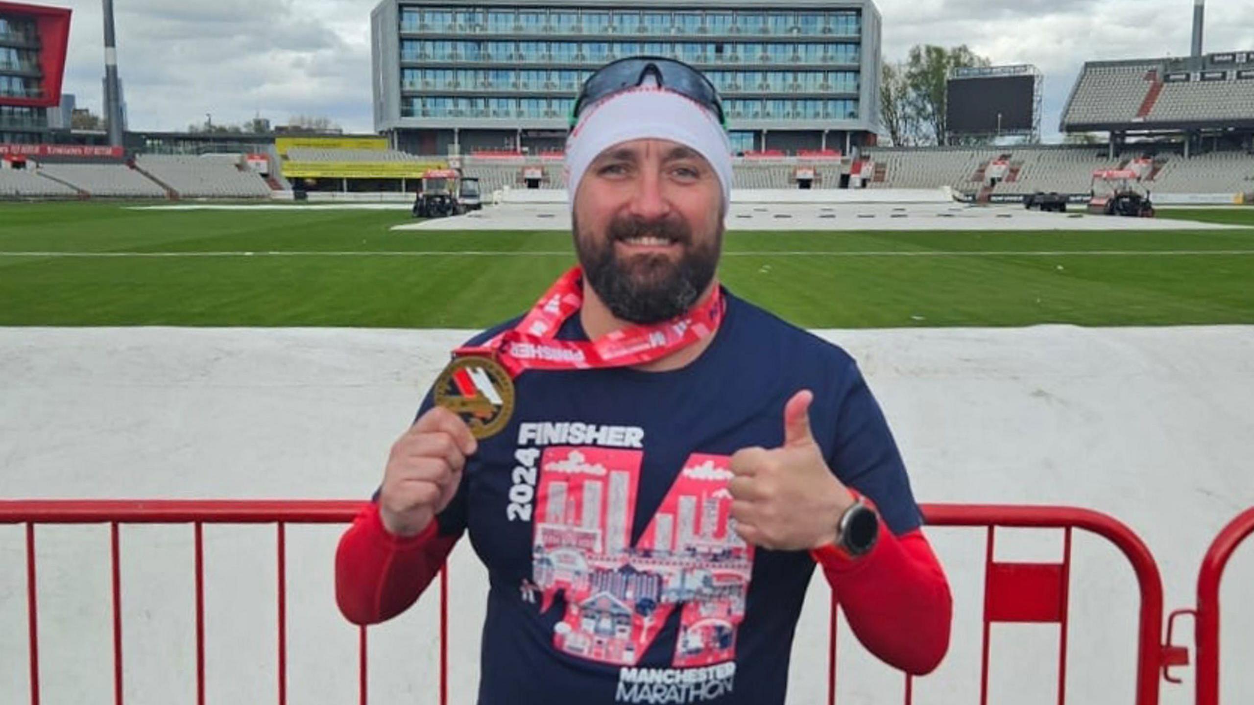 Nick Passey with dark beard wearing a white headband and blue top with his thumbs up