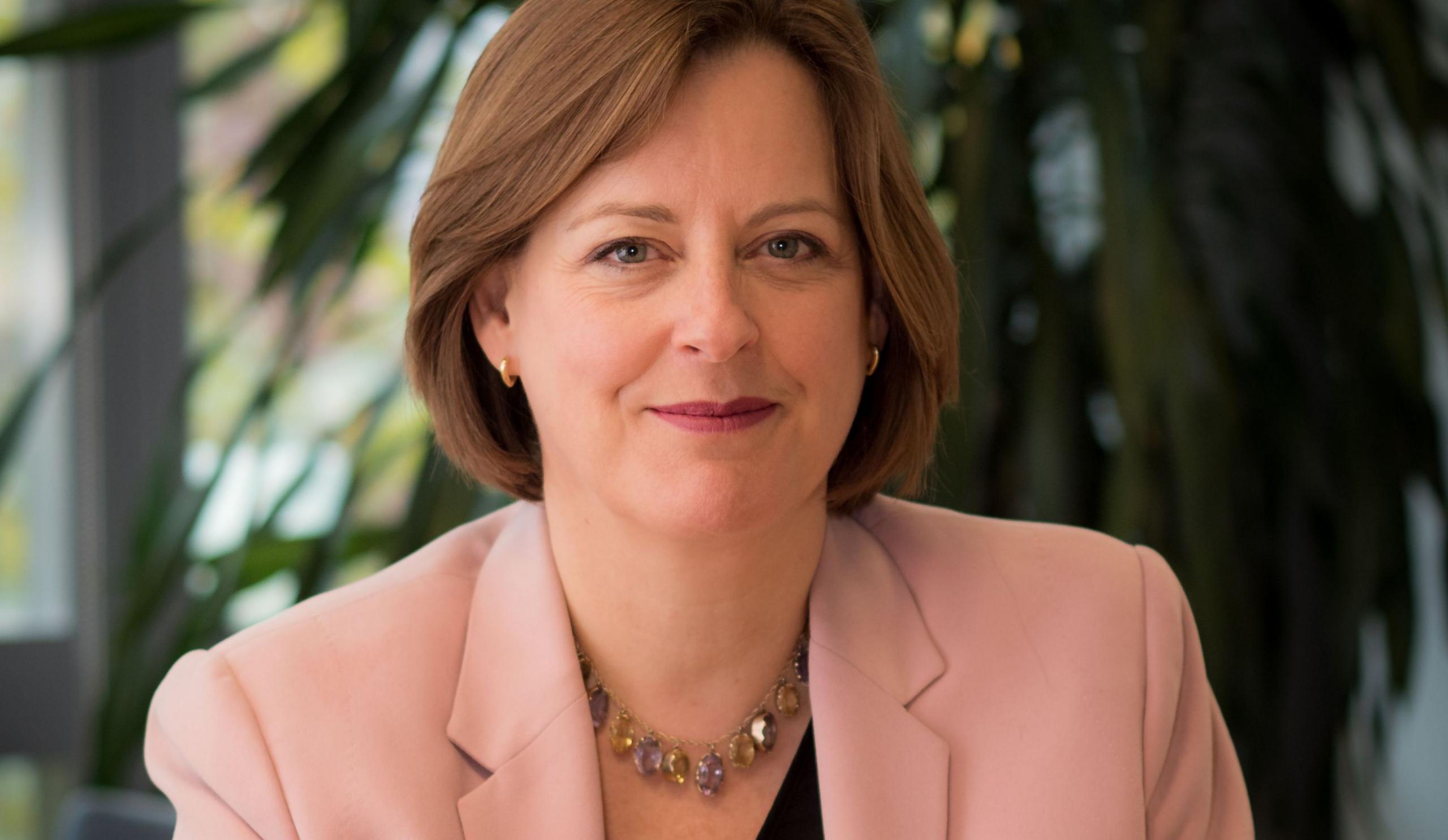 Chief Executive of Ofcom Dame Melanie Dawes smiles while looking at the camera and wearing a pink suit with a plant and a window out of focus in the background