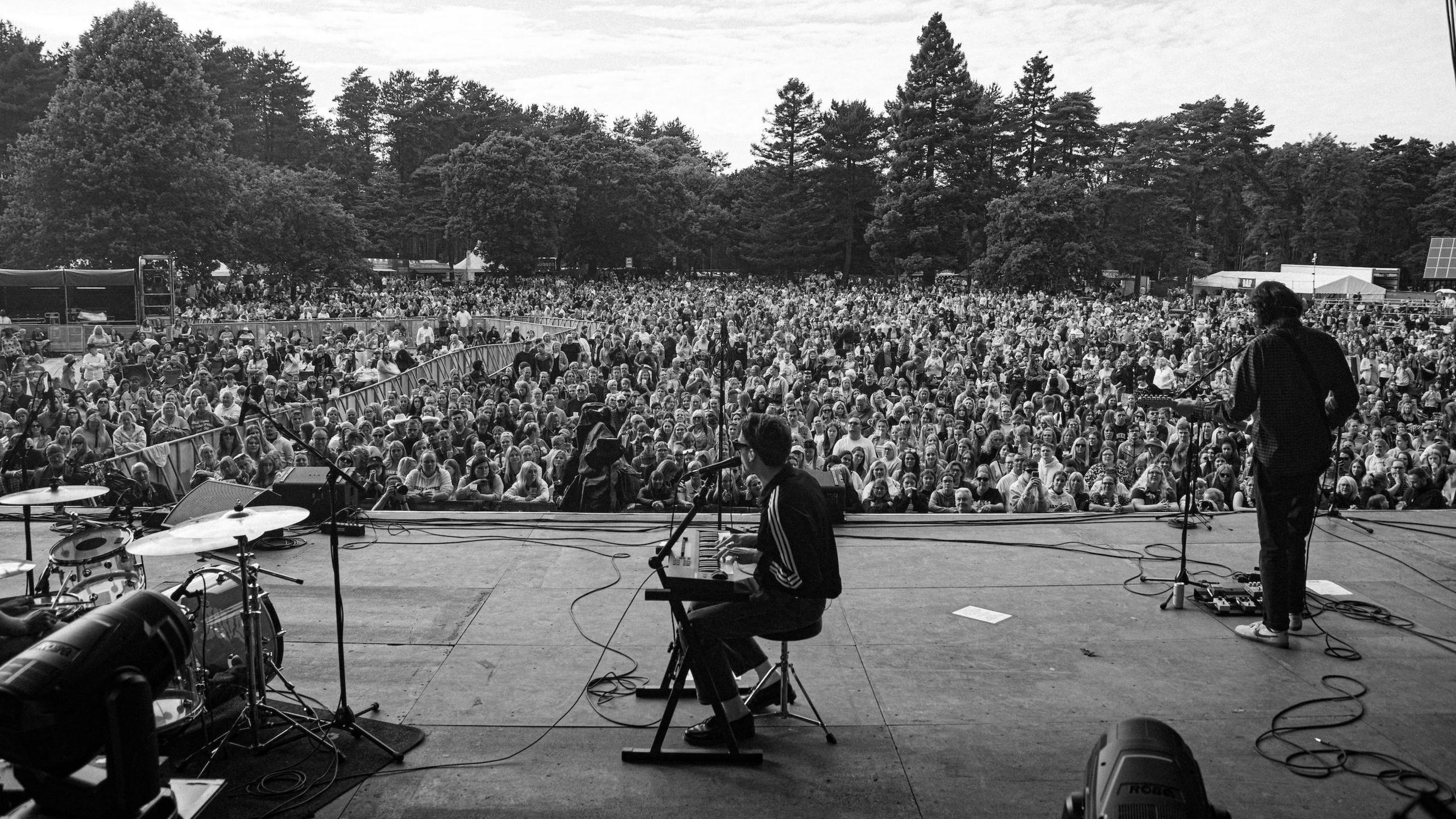 Black and white image of Devo at a keyboard in front of a large crowd and trees in the background