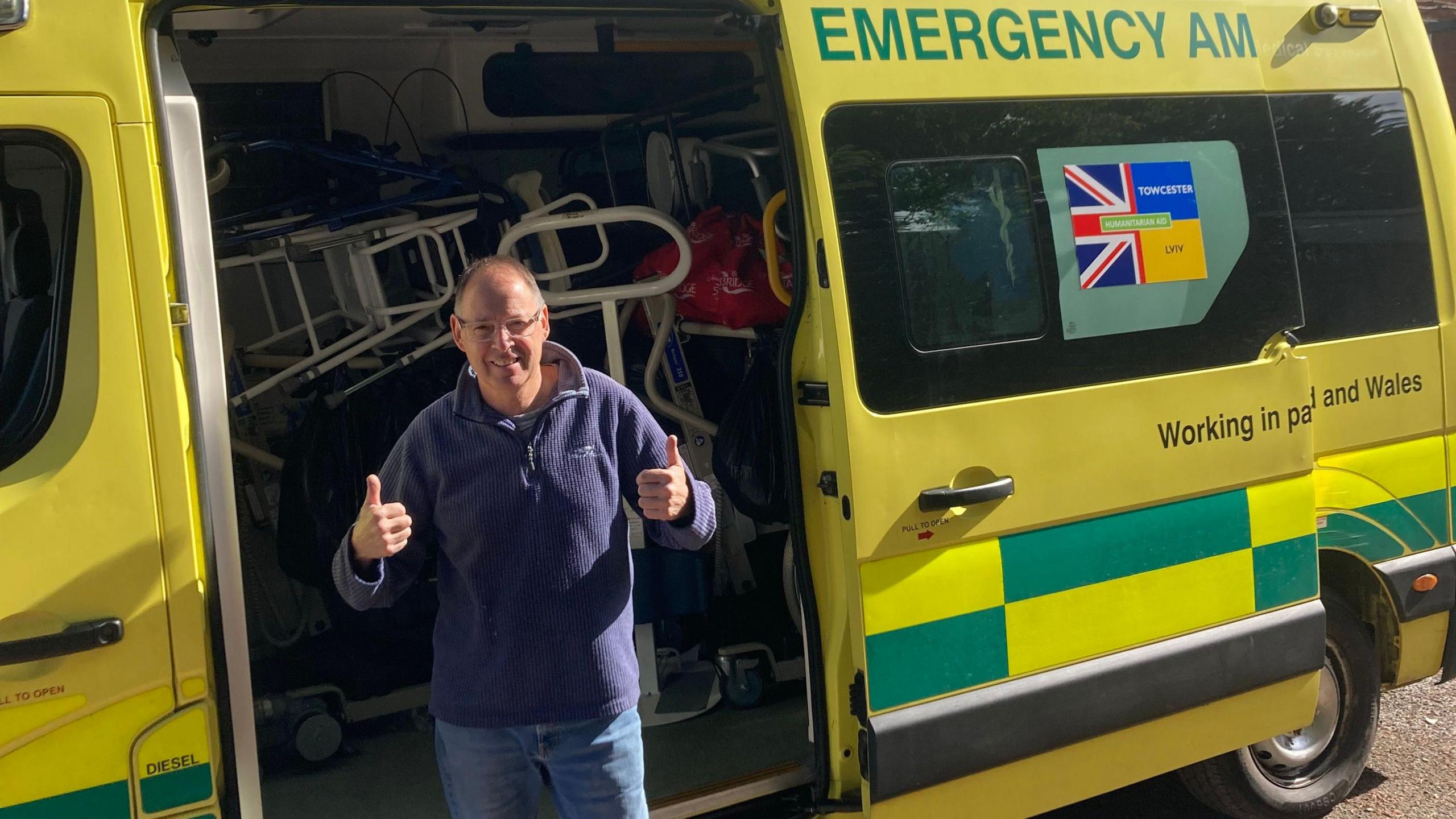 Steve Challen with short dark hair wearing a blue fleece with his thumbs up, outside the open door of an ambulance which contains walking frames and other medical items