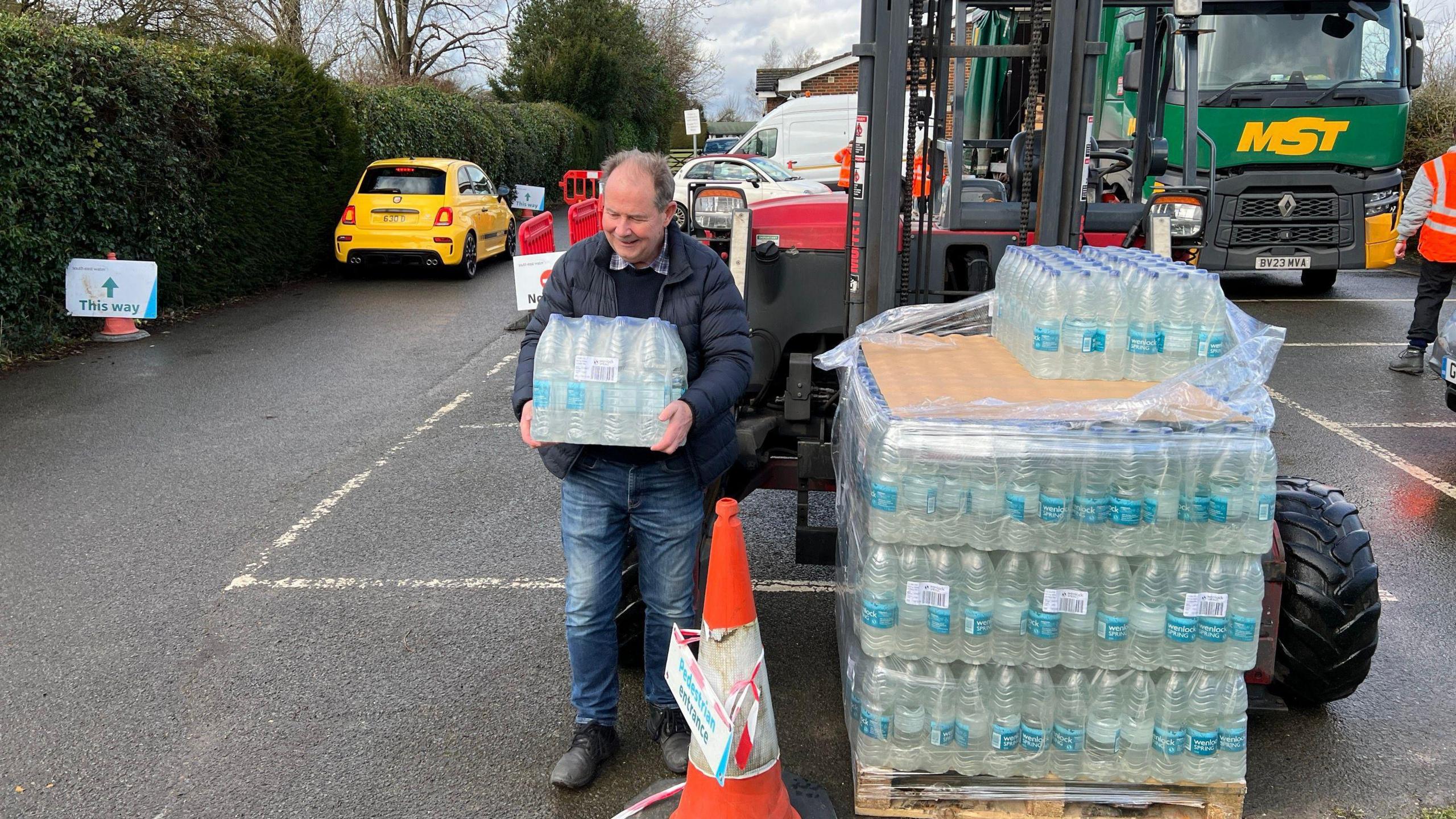 A man carrying a large pack of water bottles. There is an even larger stack of water bottles next to him on some heavy machinery, as well as vehicles in the background in what appears to be a car park. 