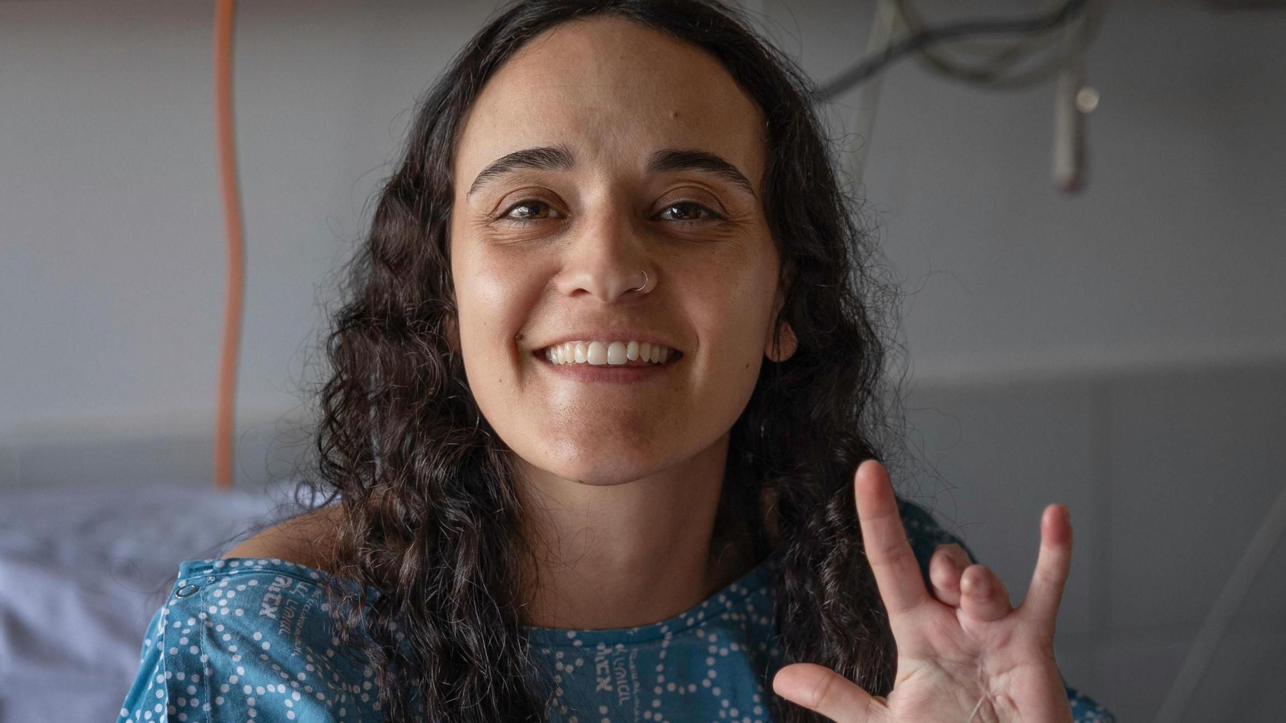 Emily Damari, a young woman with curly brown hair wearing a blue hospital gown, smiles and holds her hand to the camera while sat in a hospital bed, showing her two missing fingers.