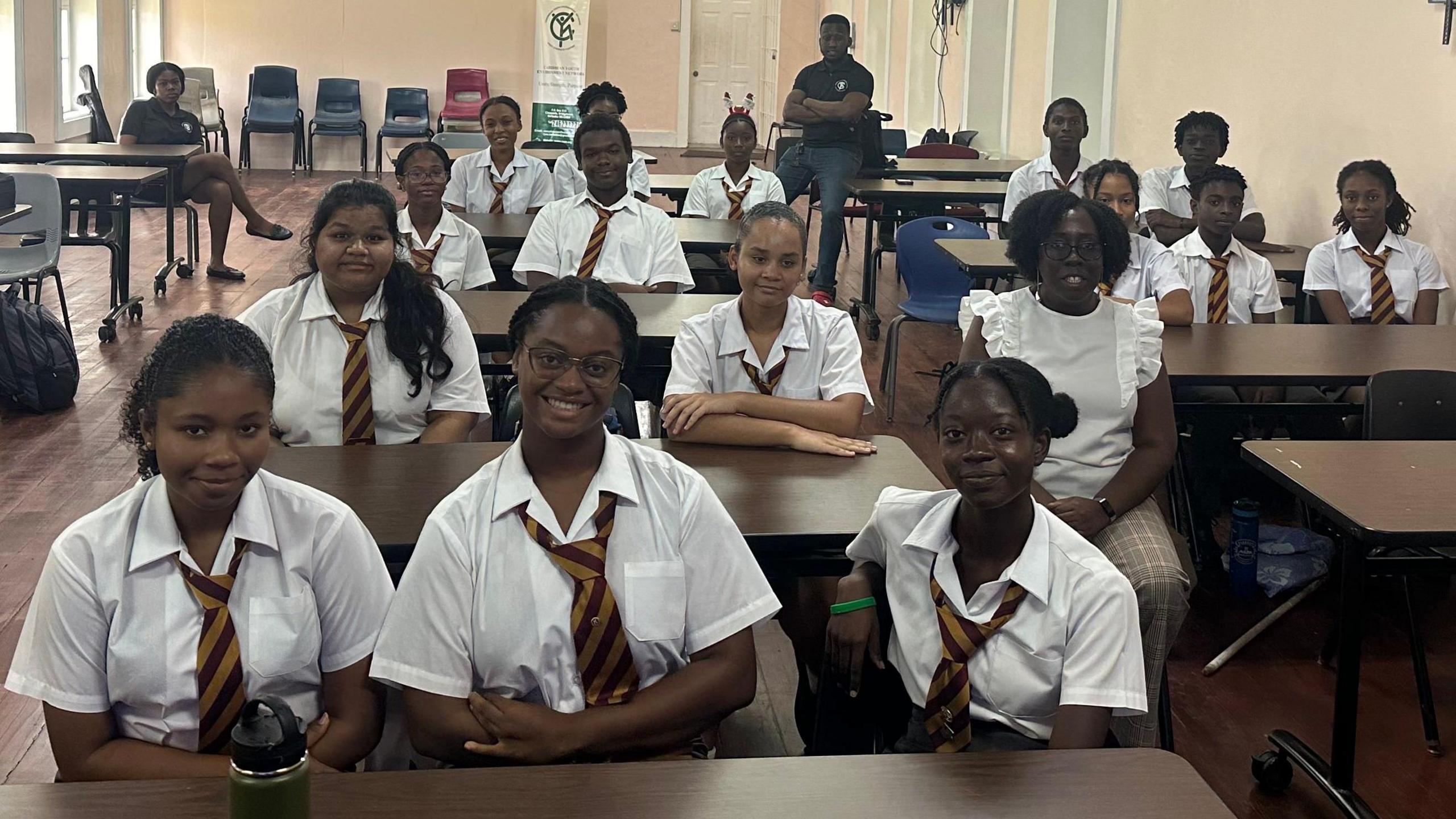 Teenage girls and boys sit behind desks in a classroom at Harrison College. All the pupils are wearing white shirts or blouses and blue-and-yellow striped ties. Two teaching staff, a man and a woman dressed in blue, are at the back of the classroom.