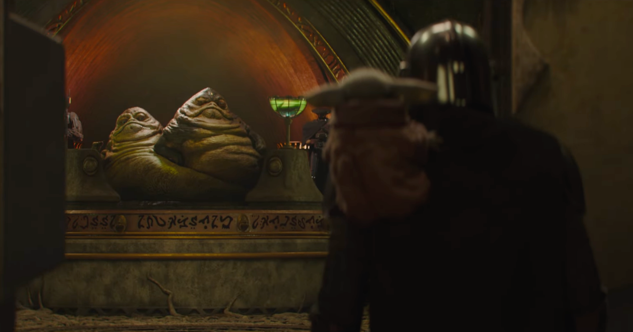 Two hutts on a throne from Srtar Wars as the Mandalorian and Grogu enter the room. The camera is positioned behind them. 