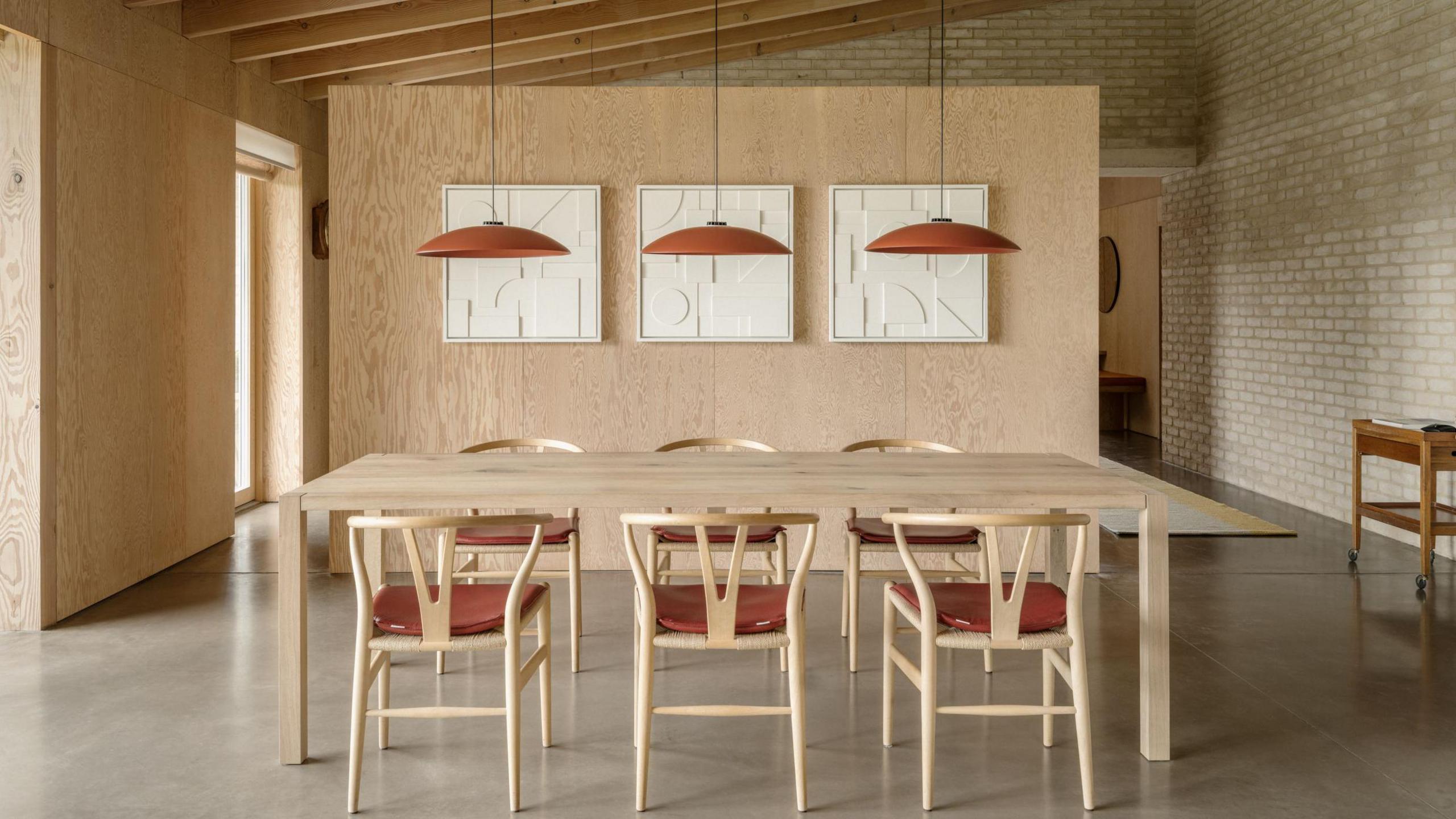 Interior of a modern house with a wood-faced wall to the left and a brick wall to the right. There is a light brown wooden table in the foreground with wooden chairs with red cushions arranged around it. There are three red lamp shades hanging over the table from the ceiling and a wooden partition is behind the table, on which three picture frames are hanging.