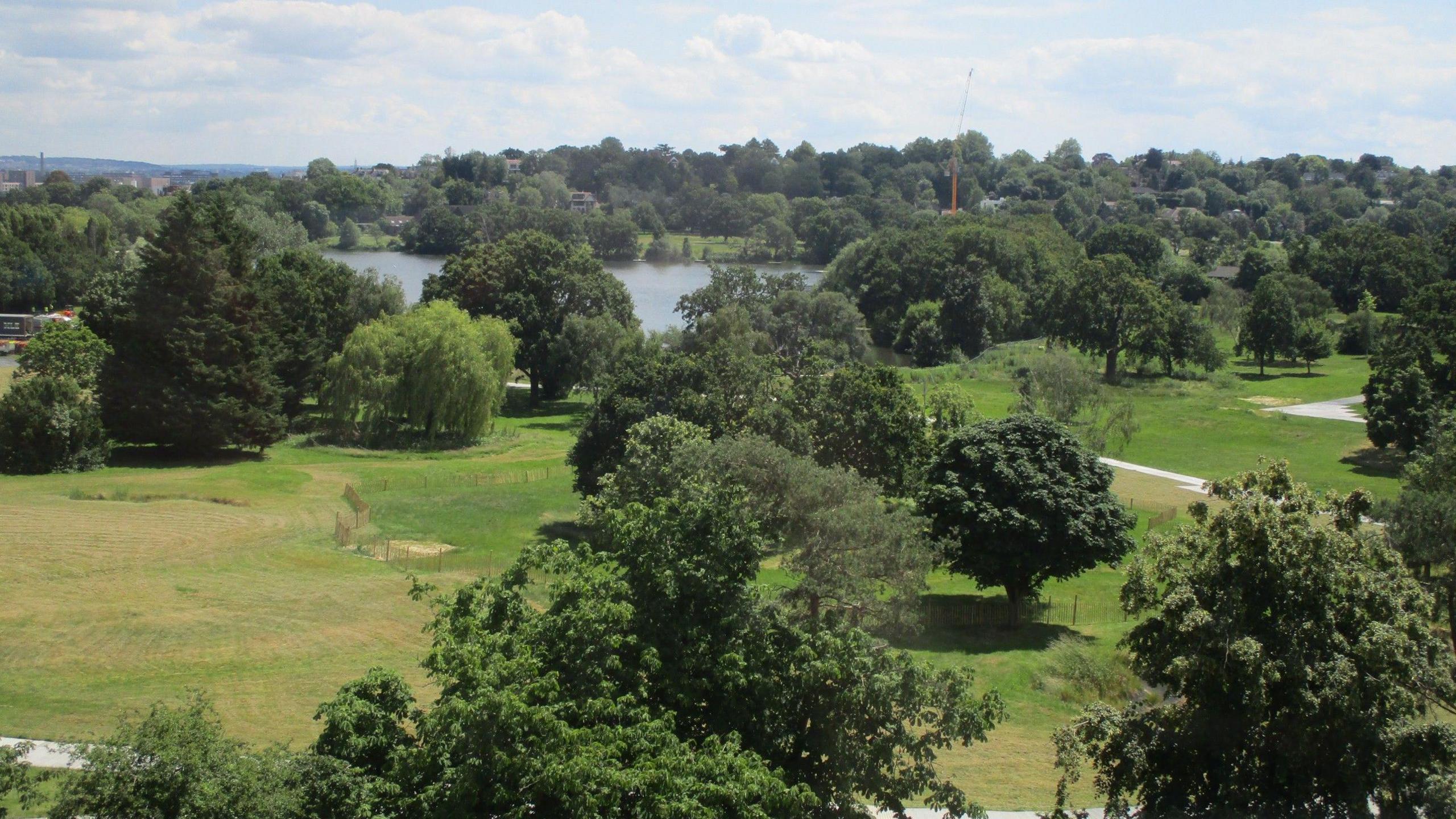 An aerial view of the current golf course within Wimbledon Park on a sunny day with blue skies. There are a large number of trees in the shot and a lake in the middle.