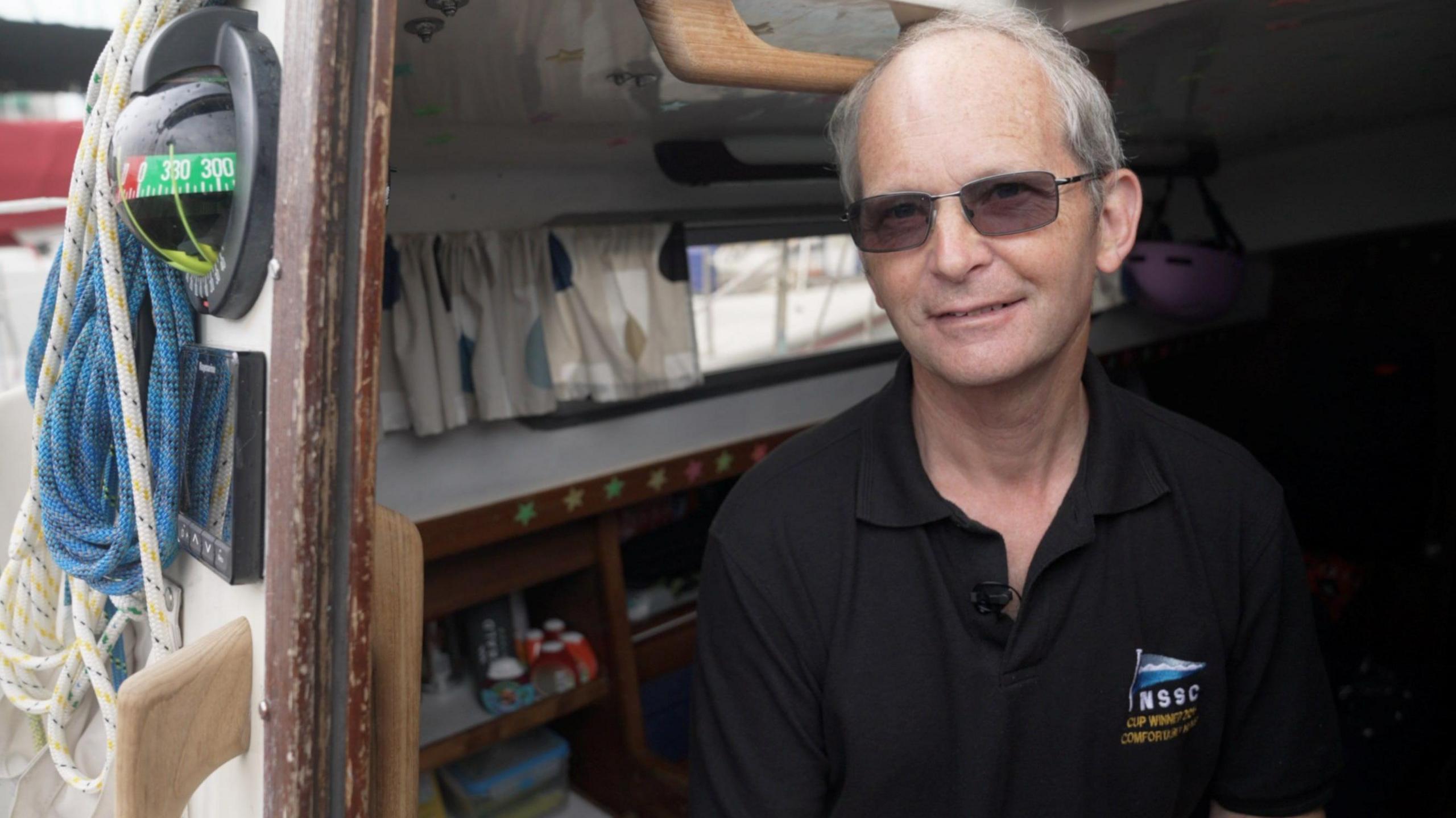 Chris sits in the cabin of the boat and looks out at the camera. There are ropes hanging on the outside. He wears dark glasses and a black t-shirt.