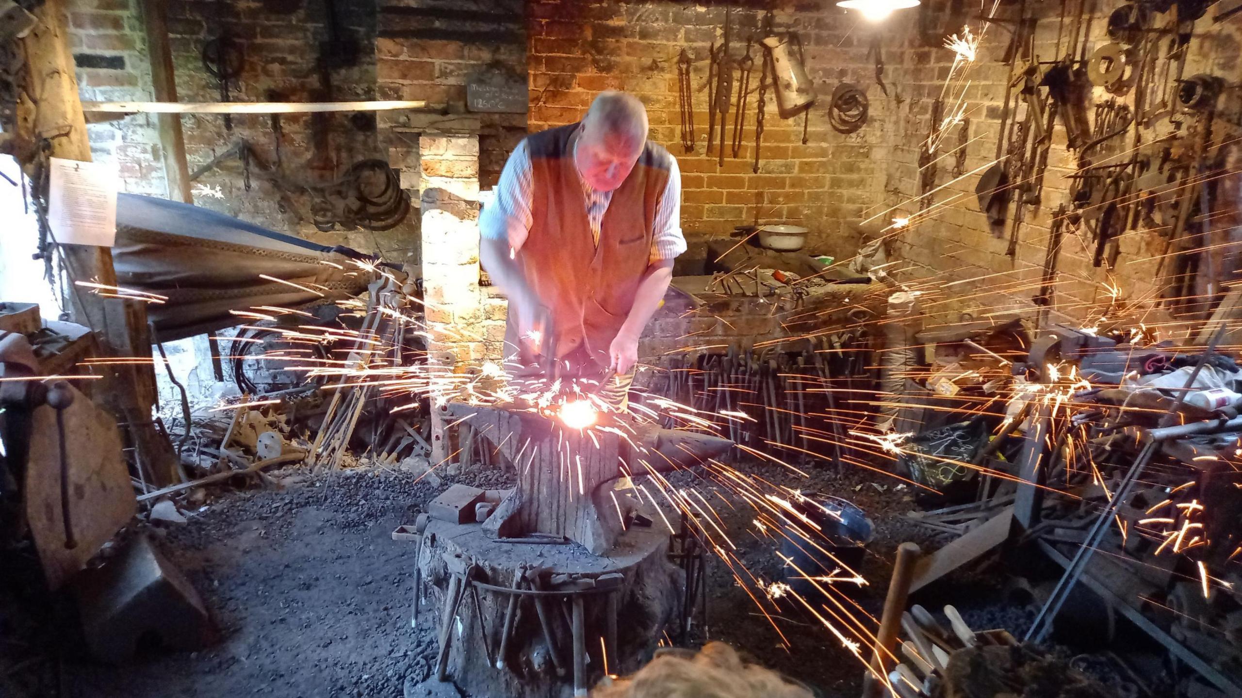 A man at work showing blacksmith skills in a workshop. His clothes include a white shirt and brown waistcoat and there are sparks around the work.