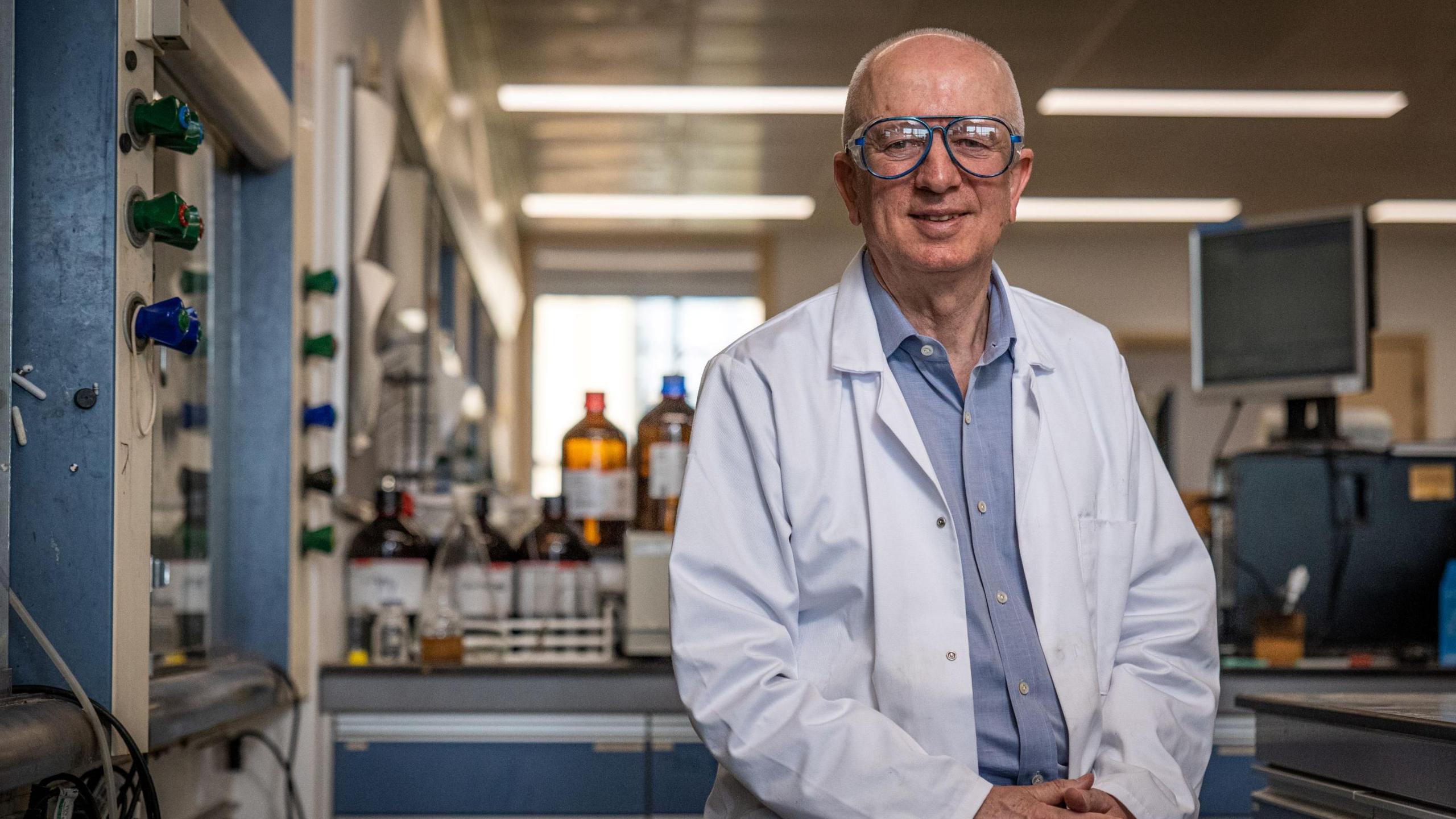 A man with large glasses on sits with his hands crossed in front of him in a laboratory setting. He is wearing a white lab coat over a light blue collared shirt.