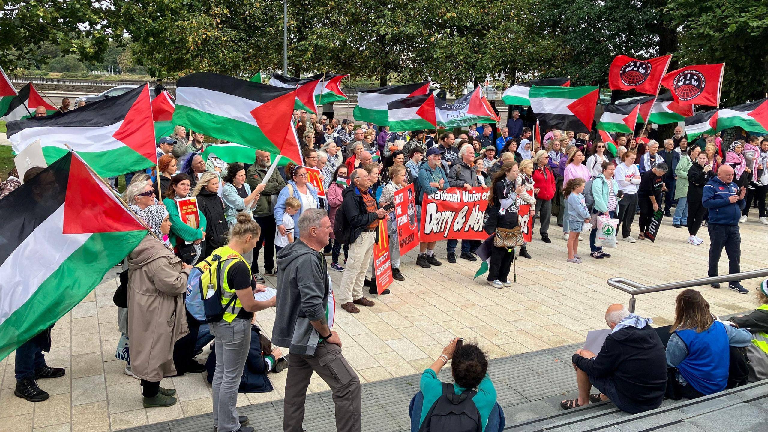 A crowd of protesters at Saturday's march in Derry. Many are carrying Palestinian flags. They are gathered on a concourse and a number of people are sitting on steps opposite, watching the demonstration