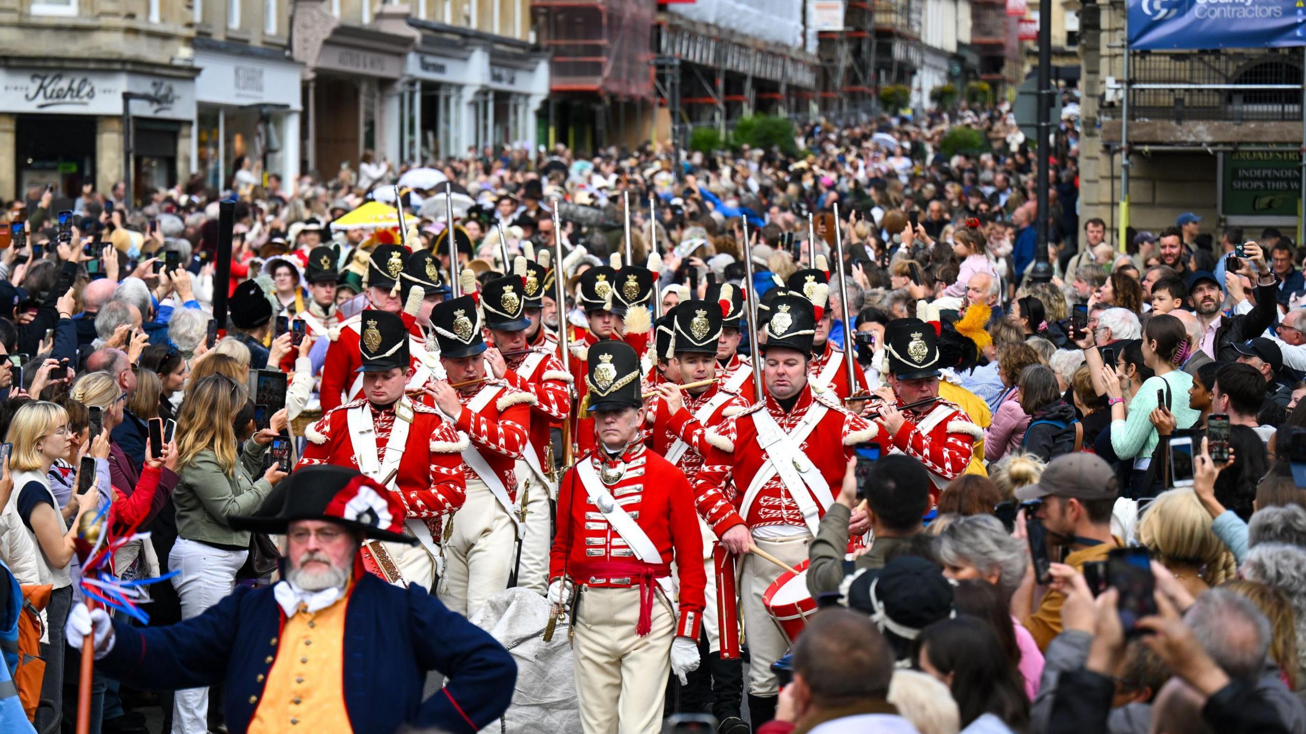 A group of men in vintage redcoat military uniforms of red jackets and white trousers march through the centre of Bath as part of the Jane Austen Festival. One of them is playing a flute while the rest have rifles held aloft. They are being led by a man in a dark blue jacket and black hat. Hundreds of people are lining the roadside, many of them taking pictures with their phones.