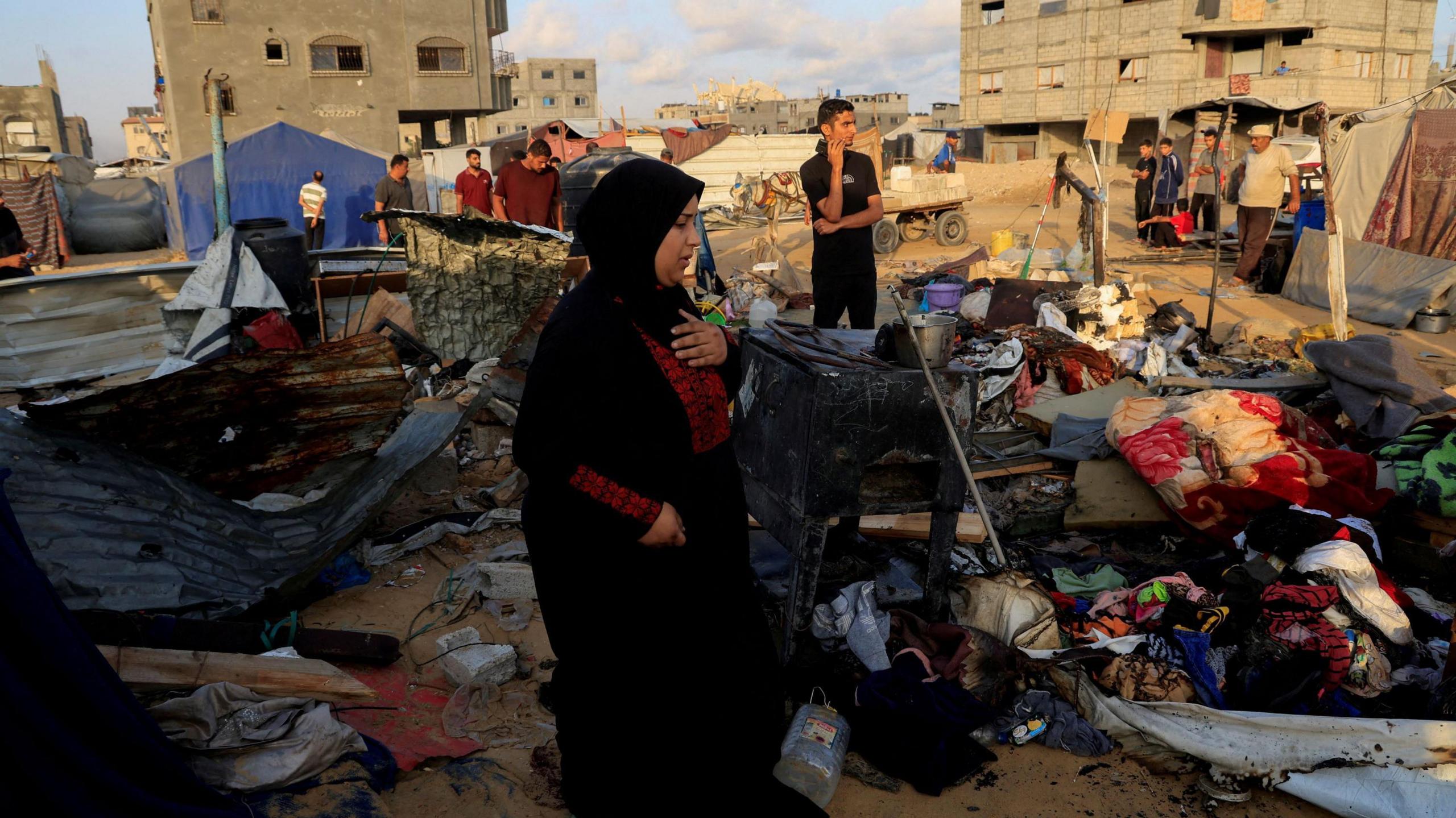 A woman wearing a black abaya and hijab holds a hand to her chest as she stands among a jumble of personal belongings - blankets, a jug, and more - after an Israeli strike.