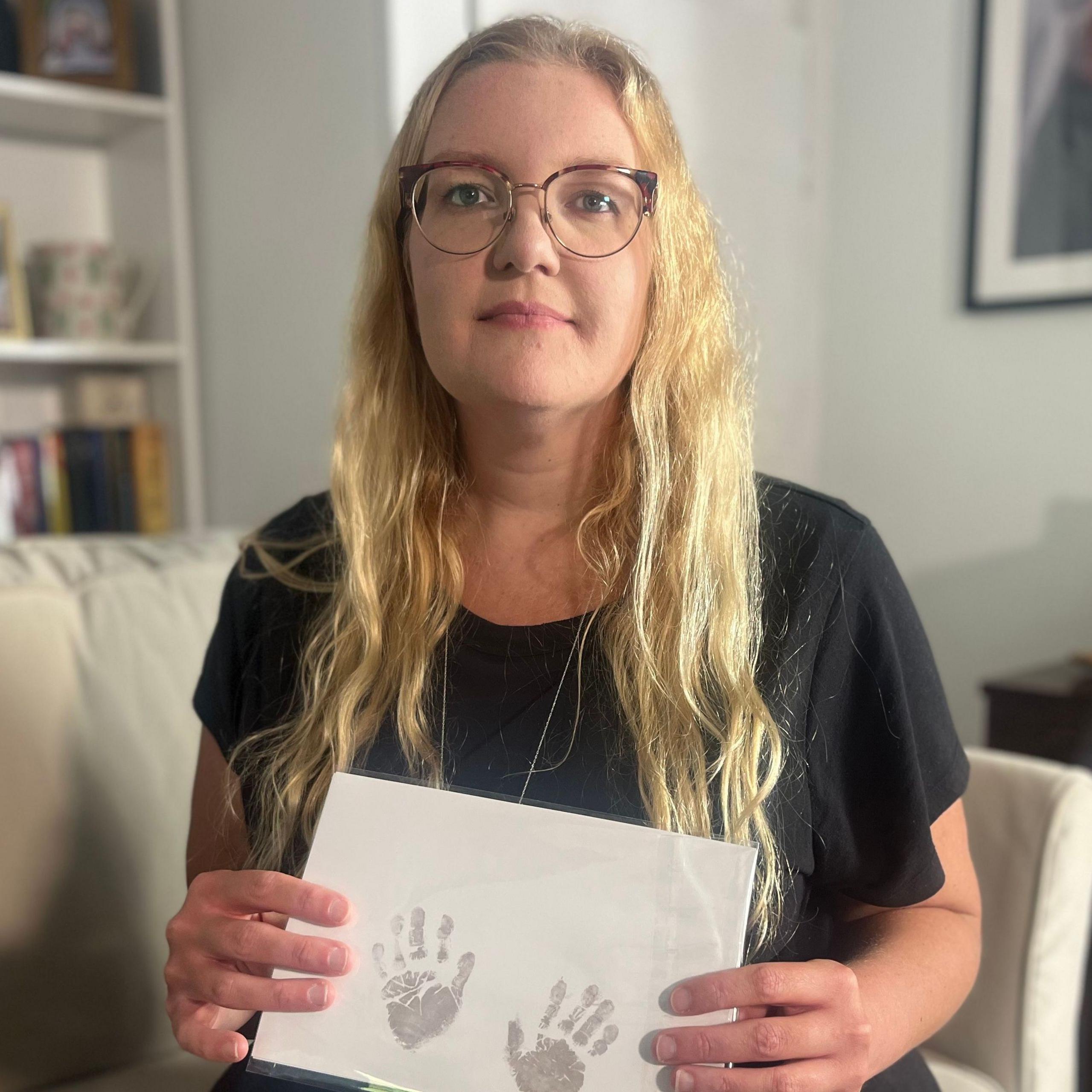 Emily Barley is sitting on a sofa in her lounge and she is holding a piece of paper which has two small handprints printed on it. Ms Barley has long blonde hair and is wearing glasses and a black top. There are framed photographs on the shelves to her right.