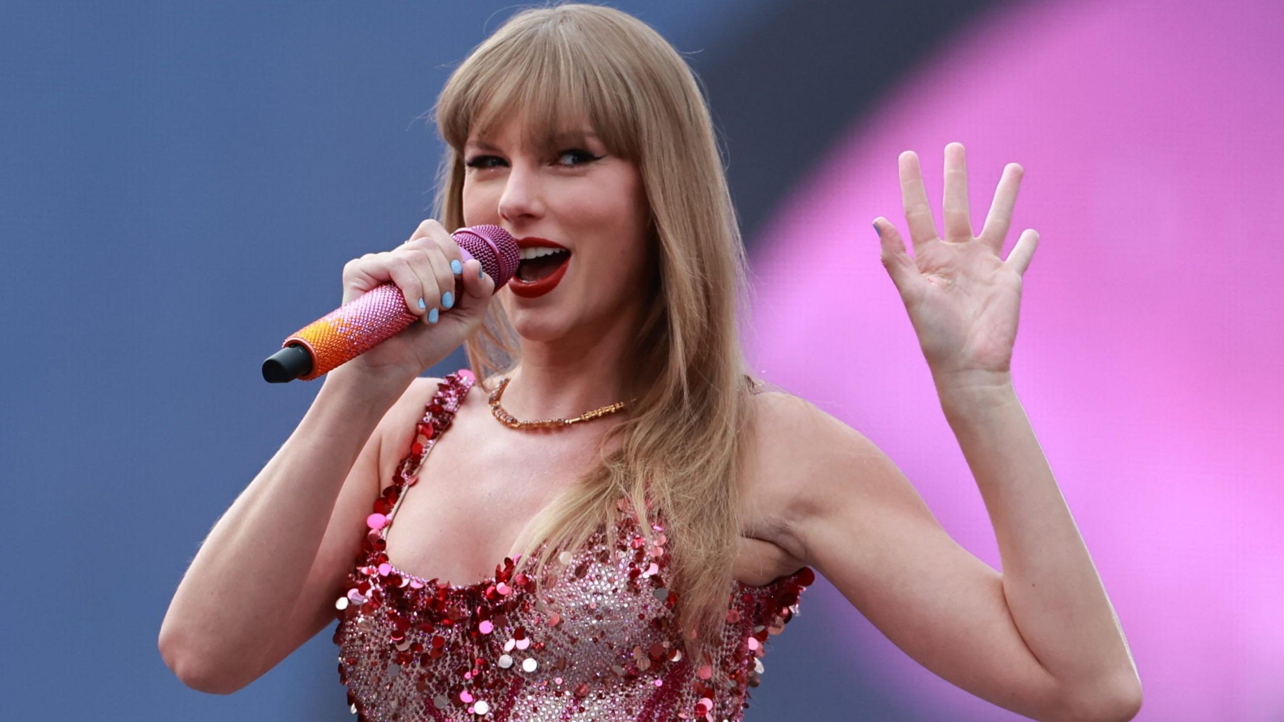 Taylor Swift, wearing a red and pink sparkling dress with a plunging neckline, holds a microphone. She is smiling and holding up her left hand.