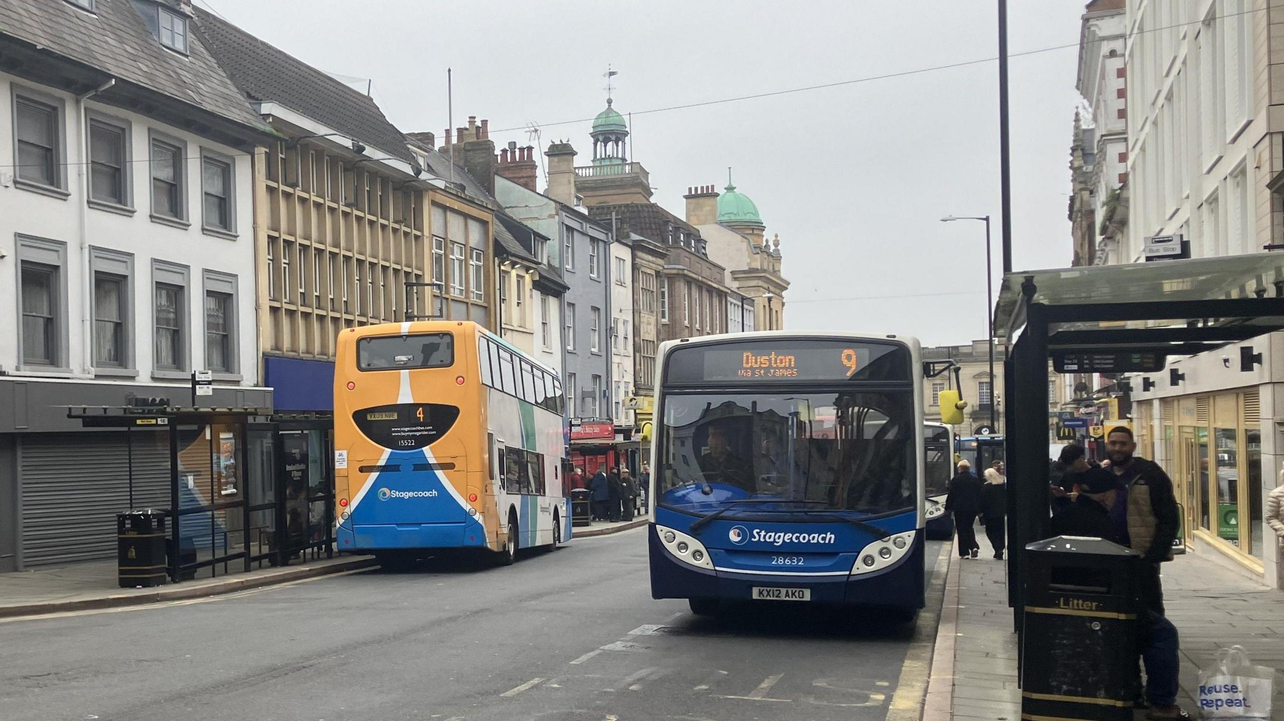  A wide shot of two buses - one blue and black Stagecoach bus with "Duston 9" on the front and the back of a yellow and blue bus. The buses are on a road with tall buildings, shops and people waiting at a bus stop.