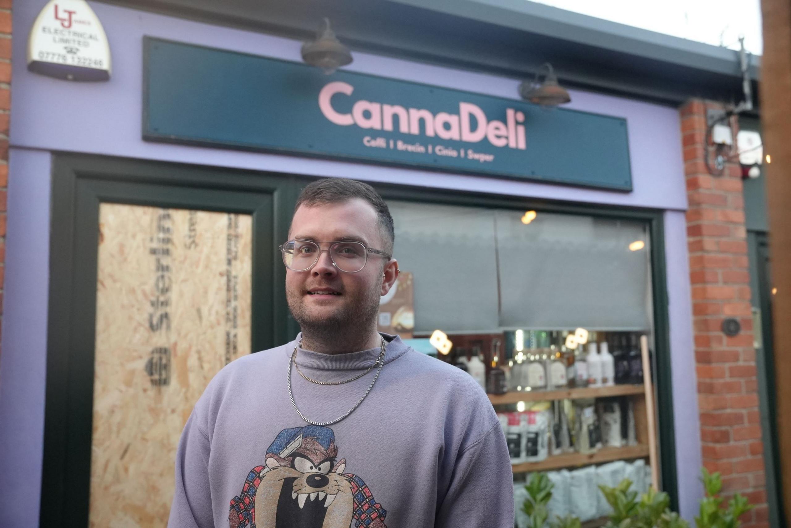 Tom Richards, owner of Canna Deli in Pontcanna, stands in front of his store. A boarded up door window can be seen in the background to the left. To his right, shelves of coffee and bottles can be seen in the window. The canna deli store sign is above his head in the background, in pink font with a navy background. 