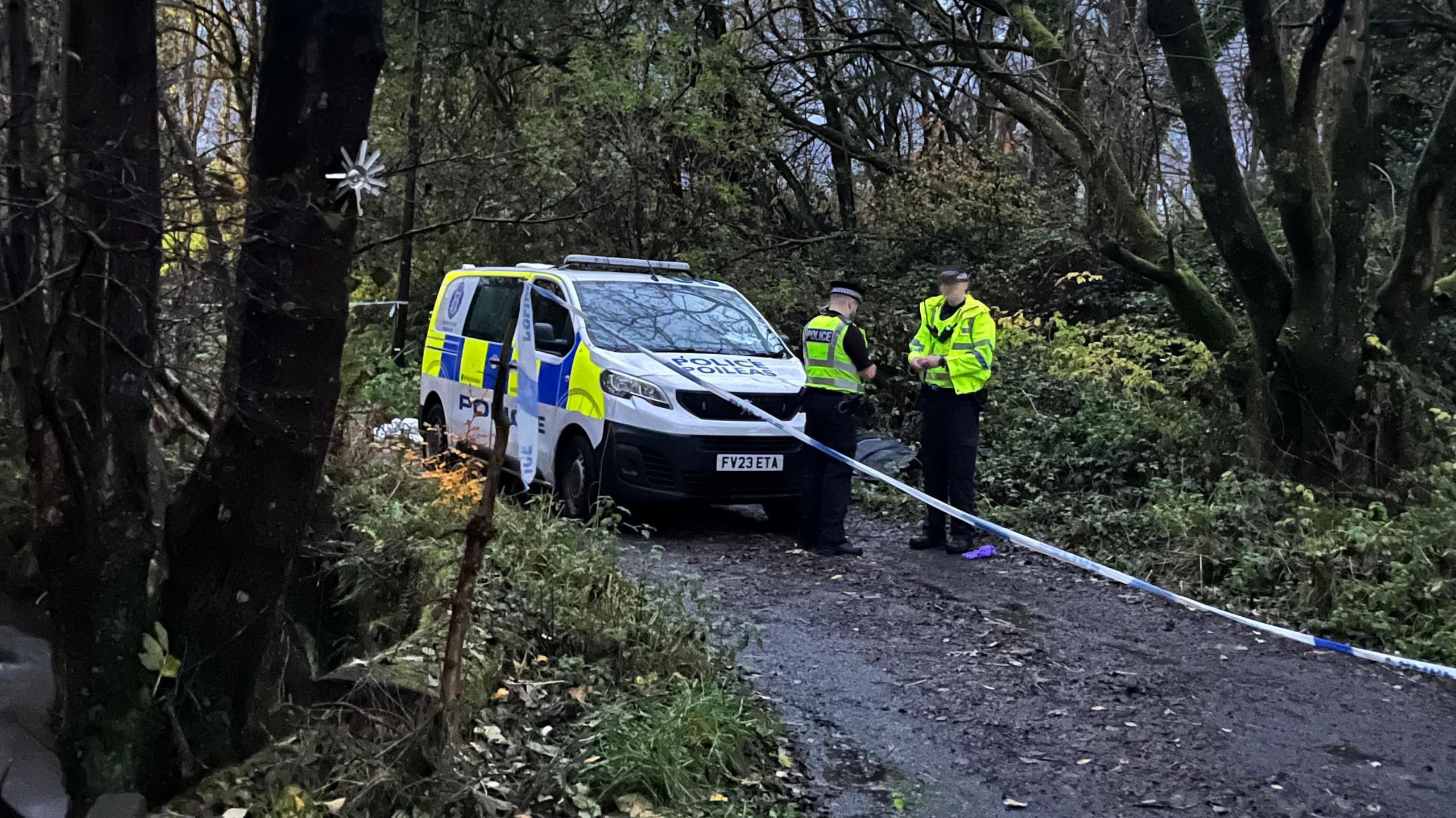 Two police officers wearing hi-vis clothing stand behind blue and white police tape in a wooded area. A marked police van is parked to their left.