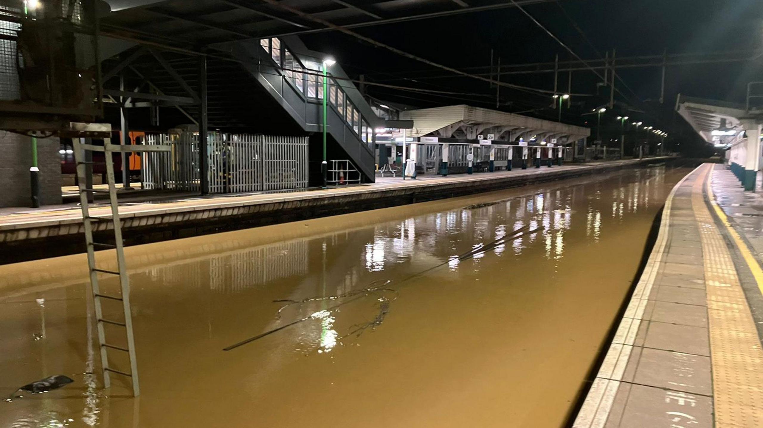 Brown flood water covering the entirety of the track at Northampton station. The water looks to be close to the platforms either side of the tracks. 