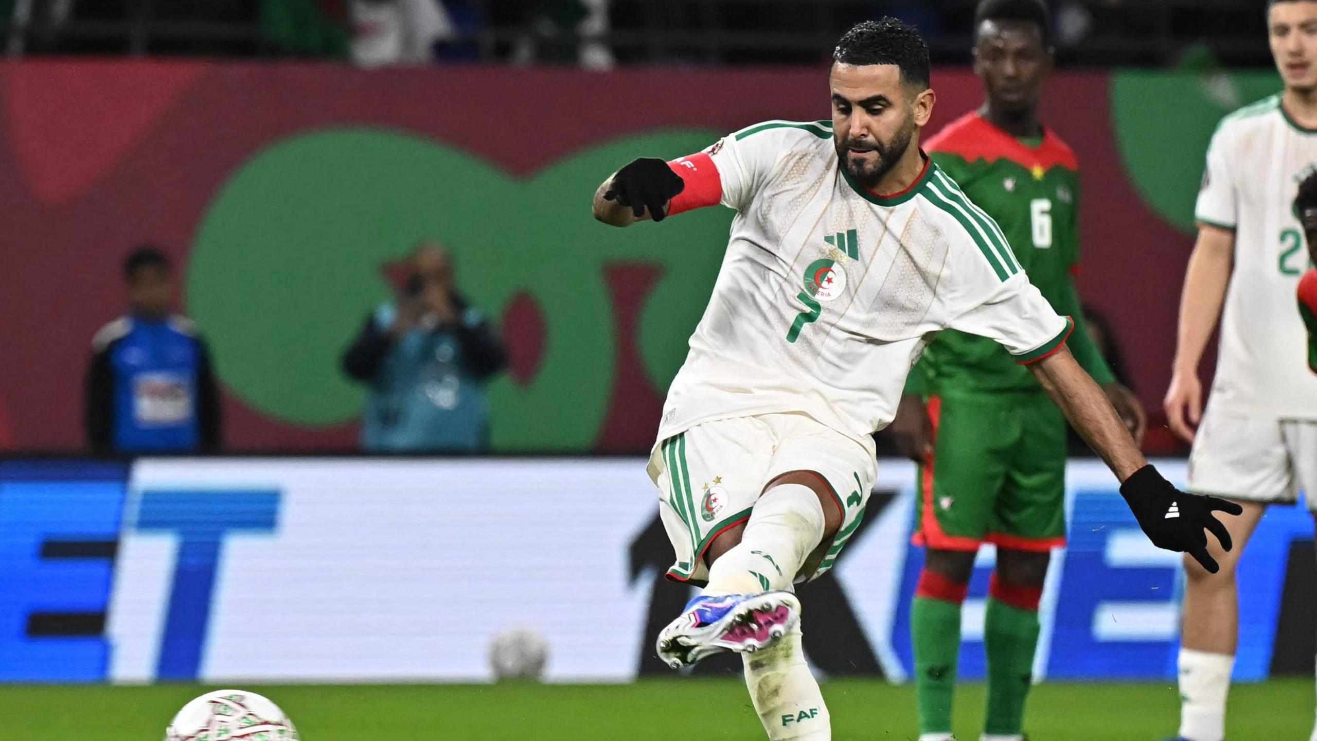 Footballer Riyad Mahrez, wearing a white jersey with green stripes and the number seven on its front, kicks the ball with his left foot while taking a penalty for Algeria against Burkina Faso at Afcon 2025. Only the top section of the ball is visible at the base of the photo as it skids along the ground after Mahrez has made a connection with it. A team-mate and an opposition player in a green kit and red trim watch on in the near background, while further away a photographer taking a picture and a ball boy are visible behind a hoarding