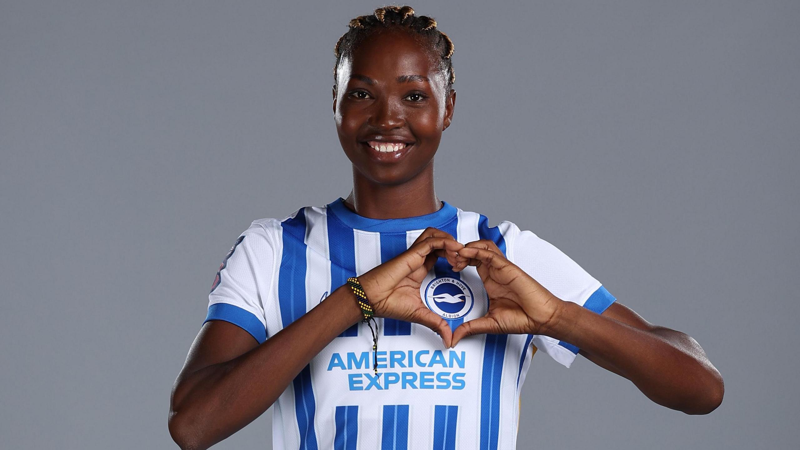 Aisha Masaka makes a heart shape over the Brighton crest while wearing a blue and white striped shirt