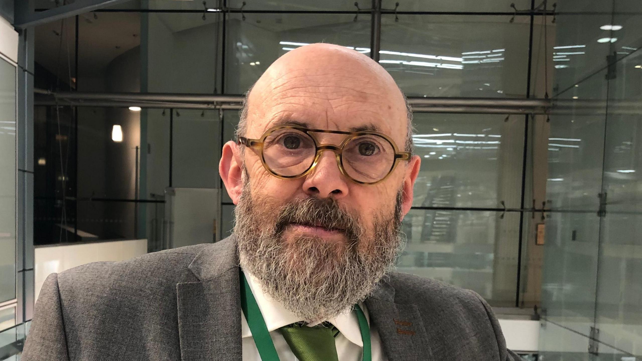 Andrew Stringer looks at the camera. He is bald with a long dark grey beard and glasses. He wears a grey suit with a green tie and a green lanyard around his neck.