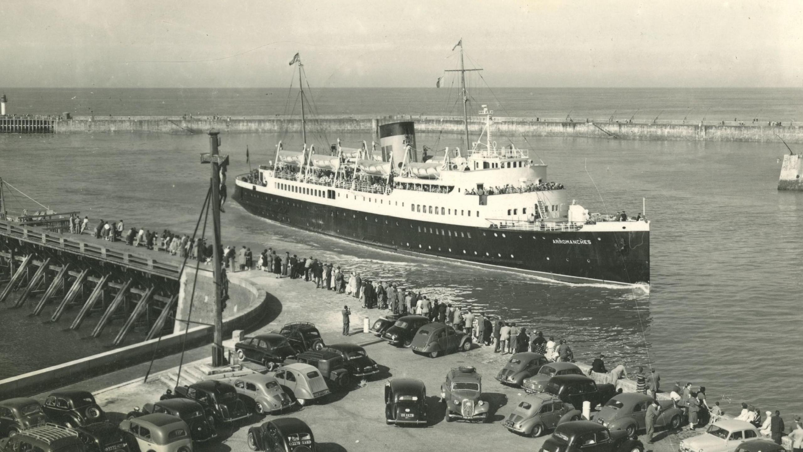 A long line of people watch as a large ferry sails close to the harbour, with many cars parked nearby