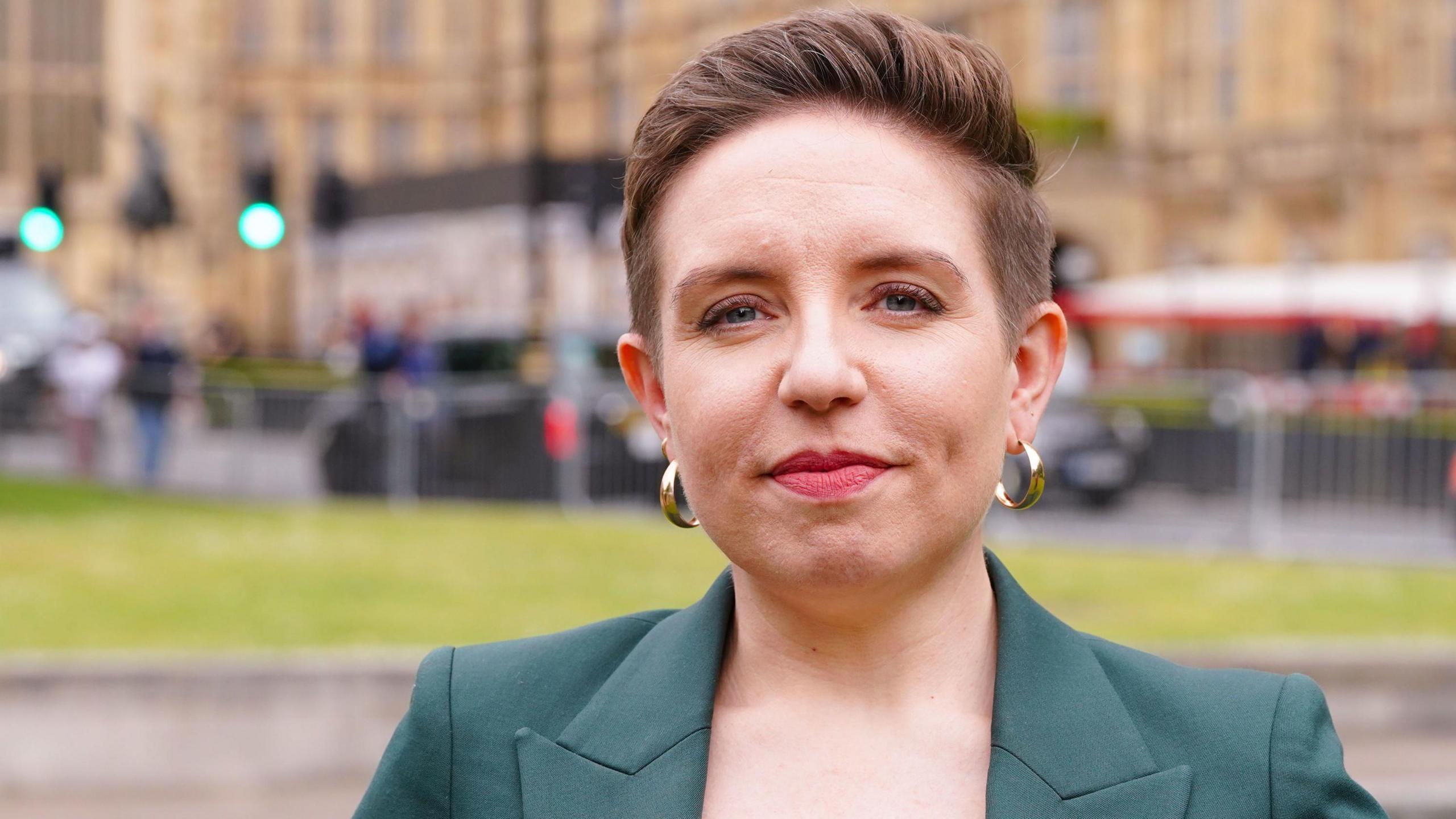 A woman with close cropped light brown hair smiles at the camera. She is wearing gold hoop earrings and a dark green jacket. The background is blurred but appears to be a grand building.