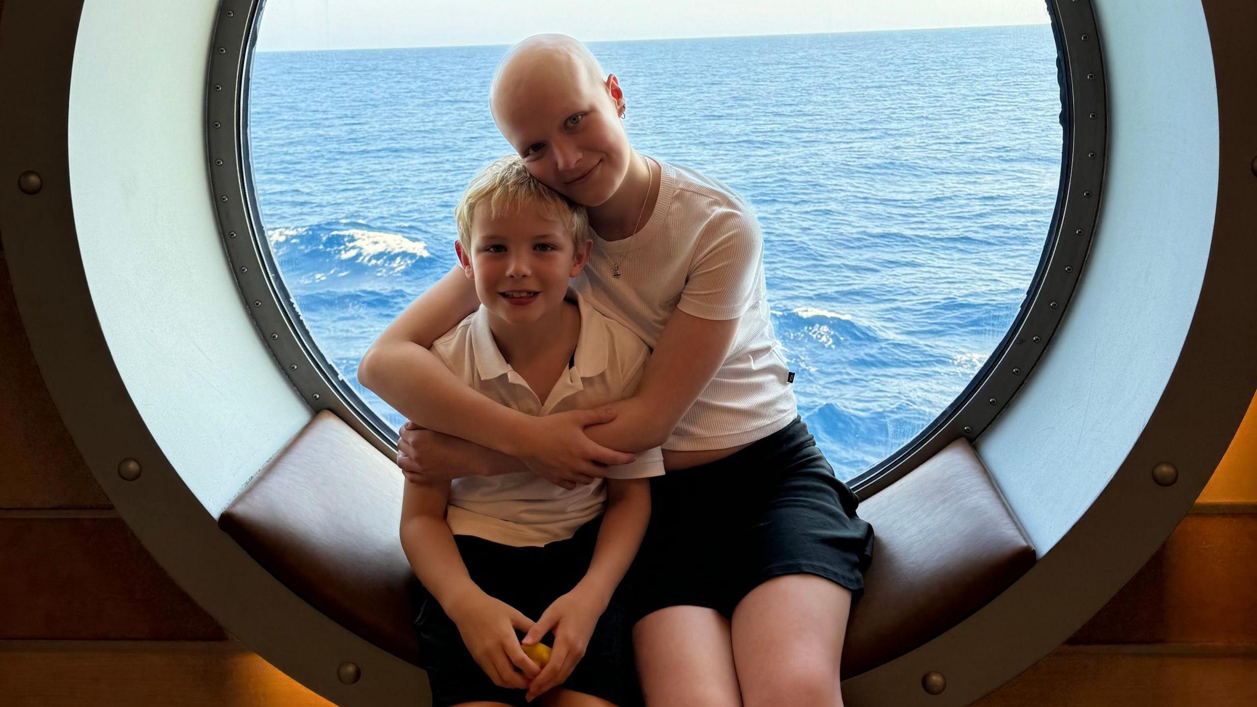 Liz hugging her brother Mateo as they sit in the window of a boat with the sea in the background. 