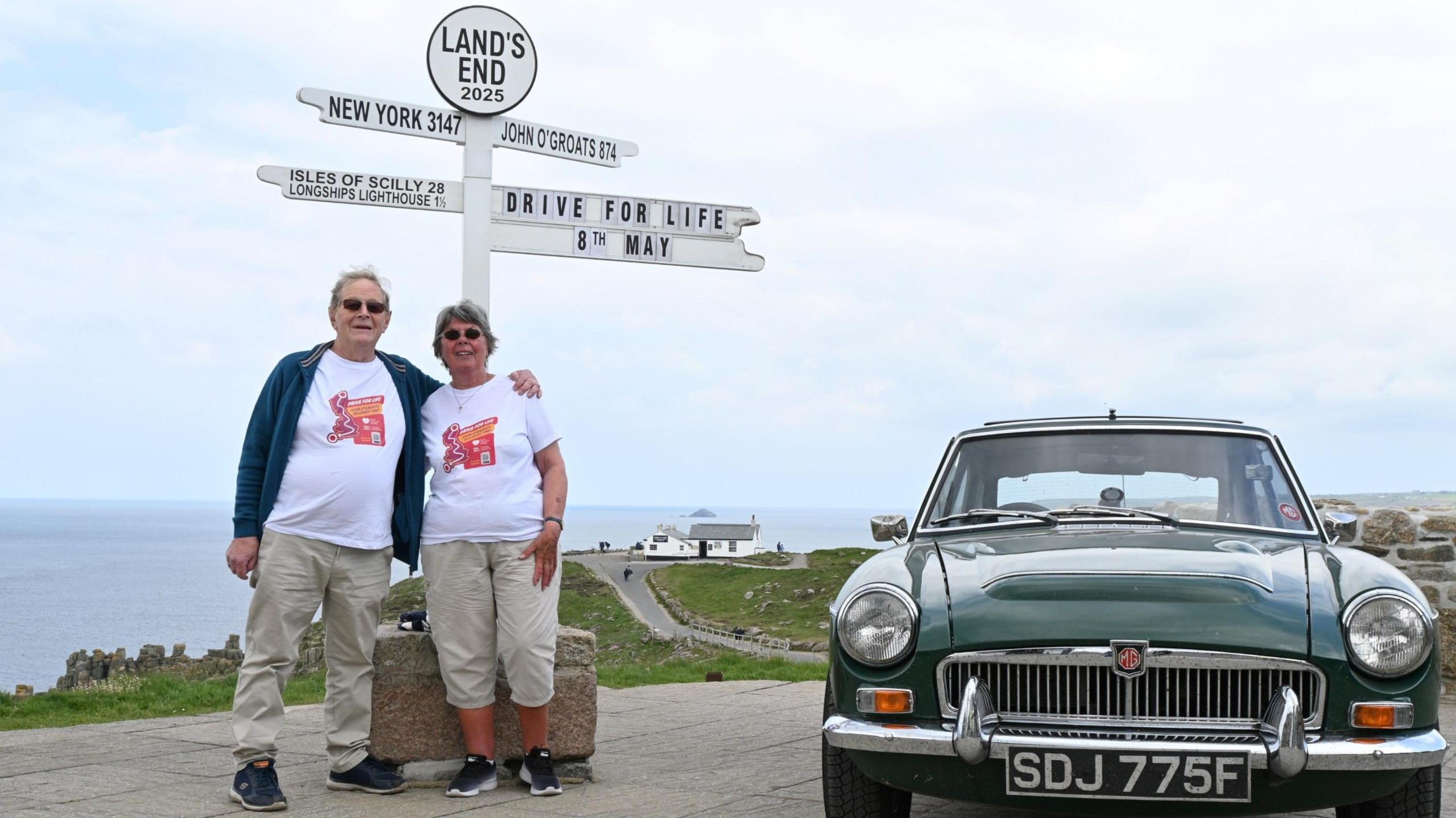 A couple standing under the Land's End finger post and next to a classing car in British Racing Green
