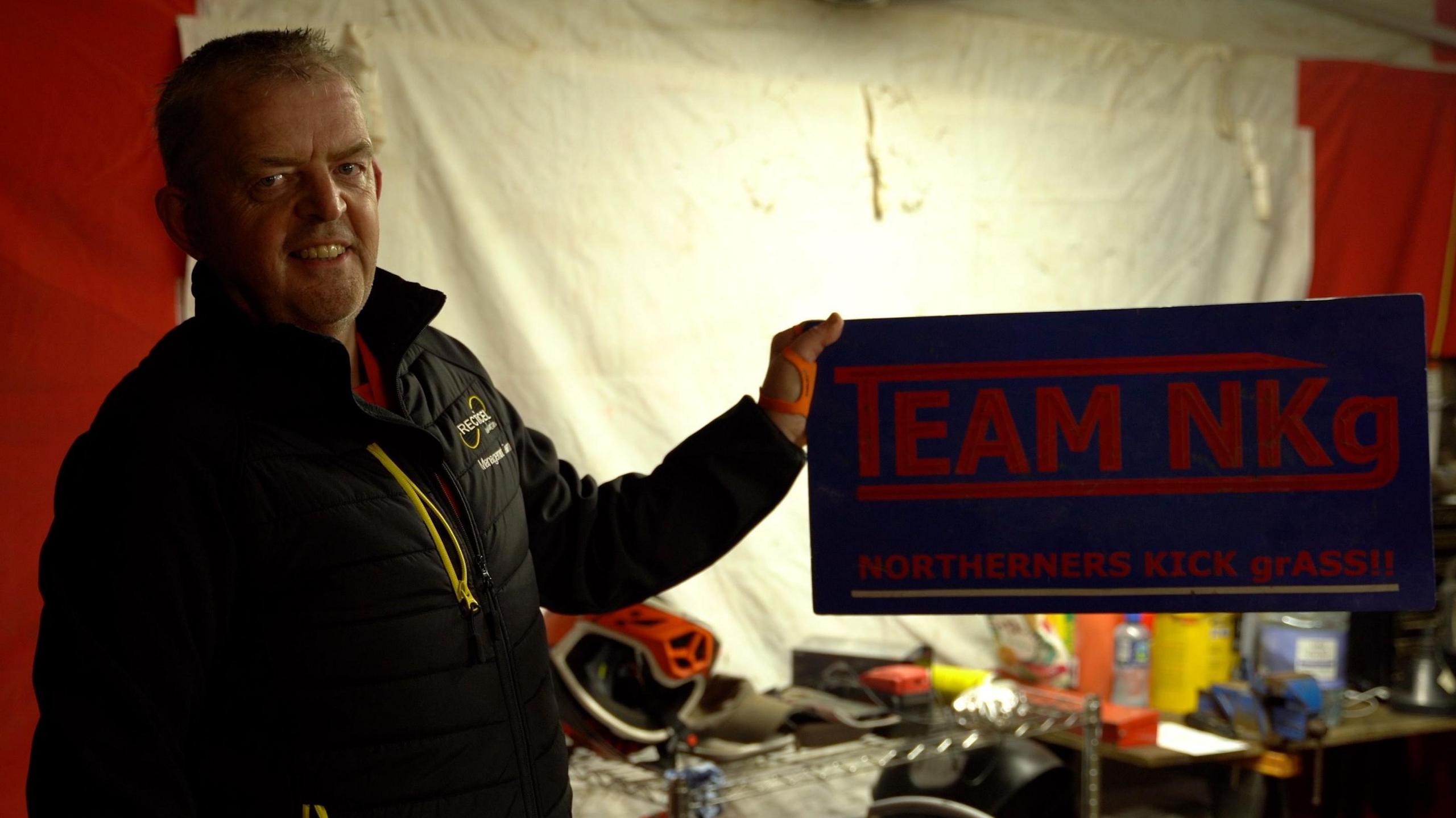 A man is holding a sign that reads "Team NKG: Northerners Cut Grass"