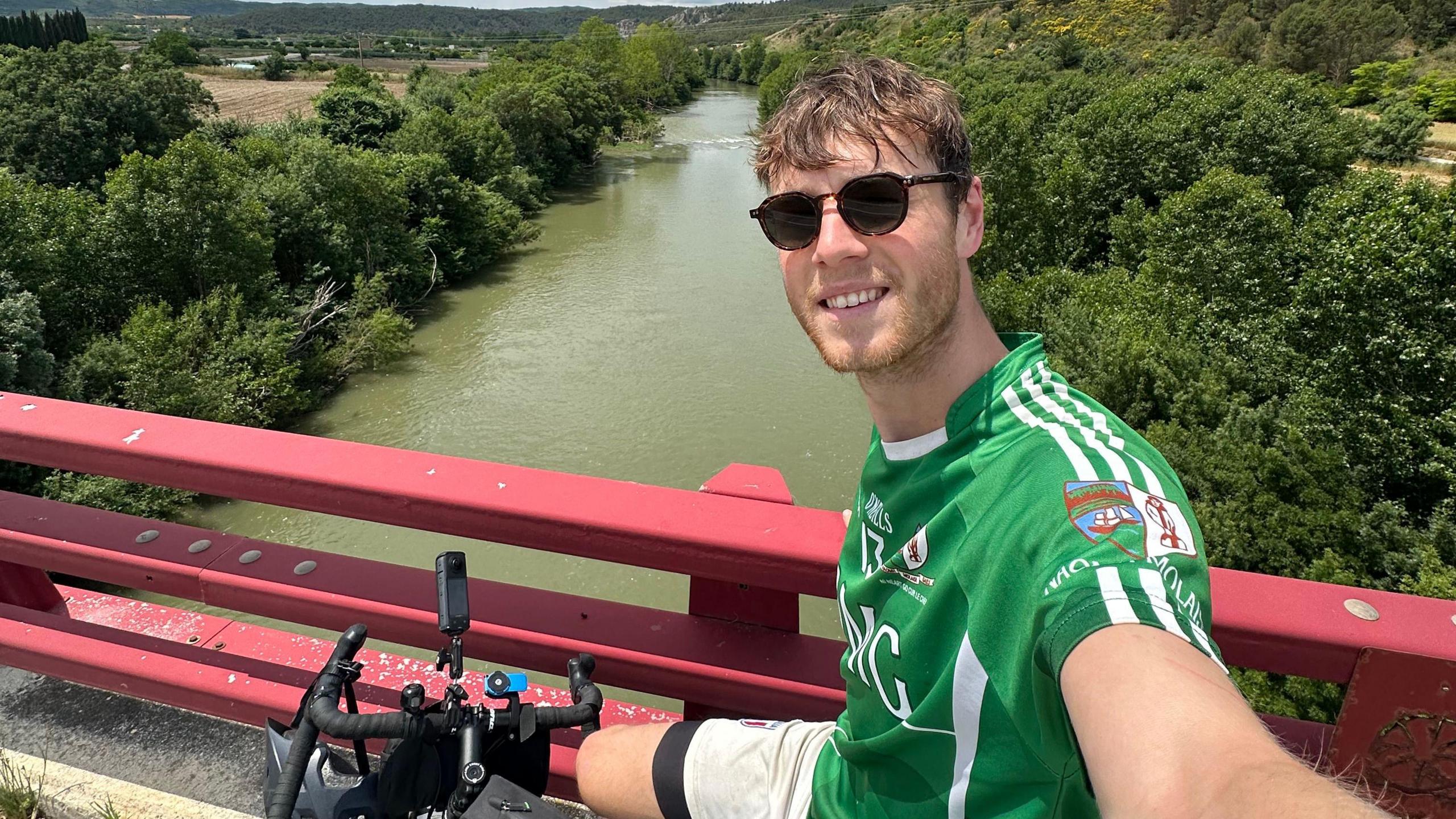 Mr White takes a selfie from his bike, which is positioned by some red railings on a bridge over a large river. He is wearing a green sports top and dark sunglasses.