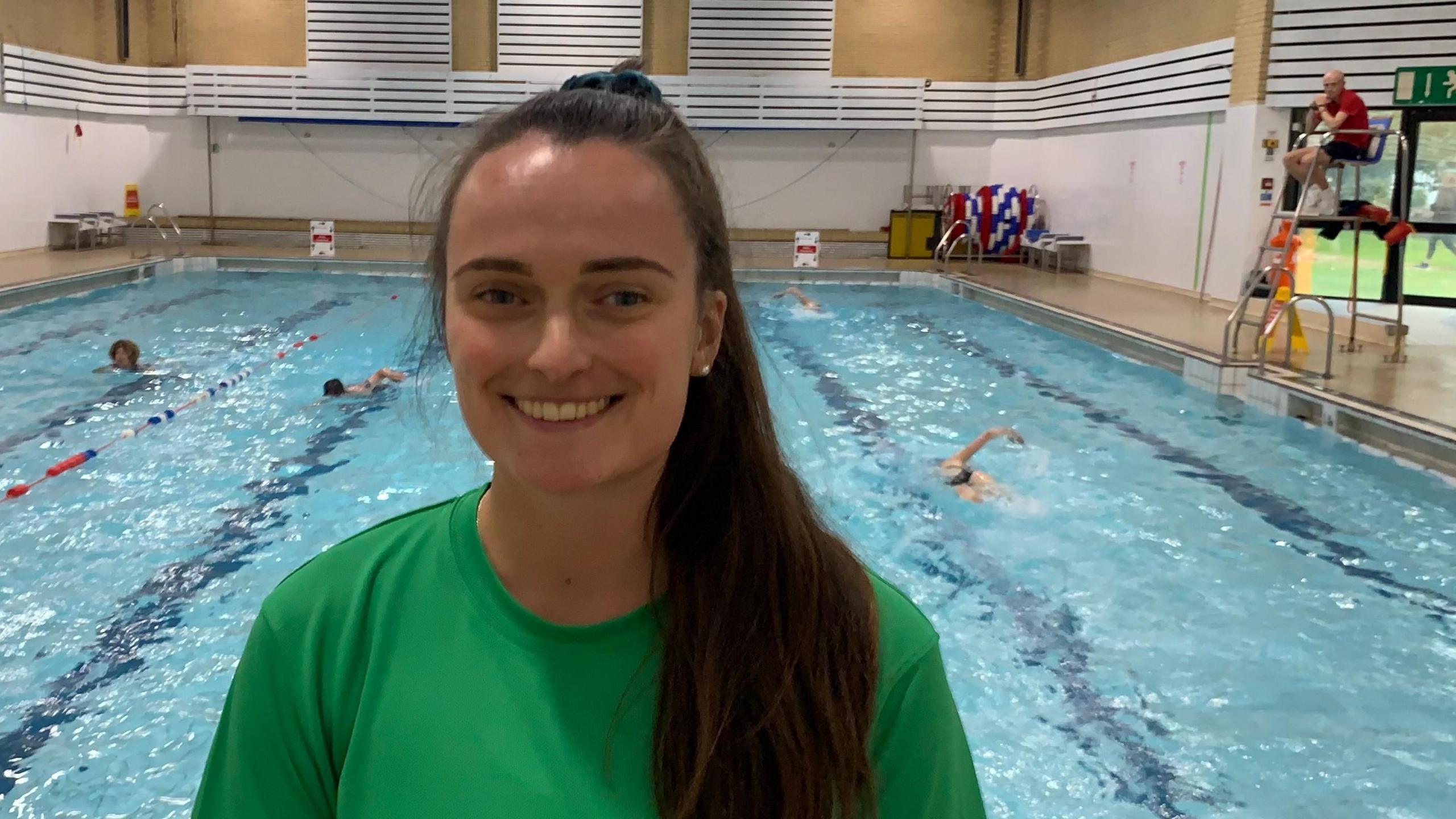 Danii Vipond Houghton is wearing a green top and is standing in front of the swimming pool at Kendal Leisure Centre. People are swimming in lanes in the pool behind her. A lifeguard is sitting on a high seat on metal stilts.