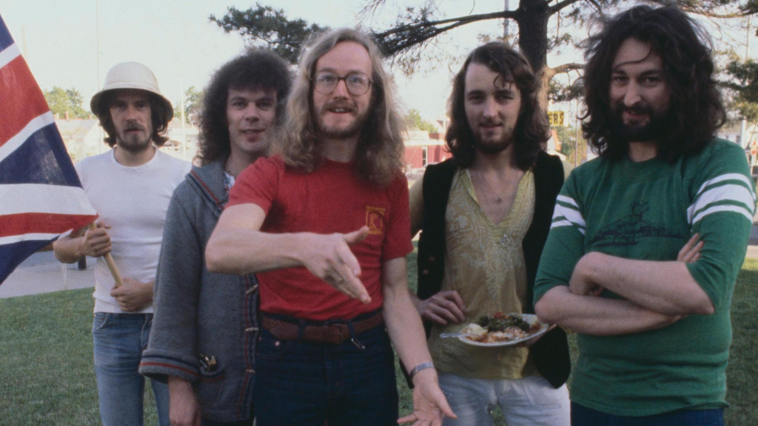 The five members of Supertramp standing together outdoors in a row, with bassist Dougie Thomson holding a union flag