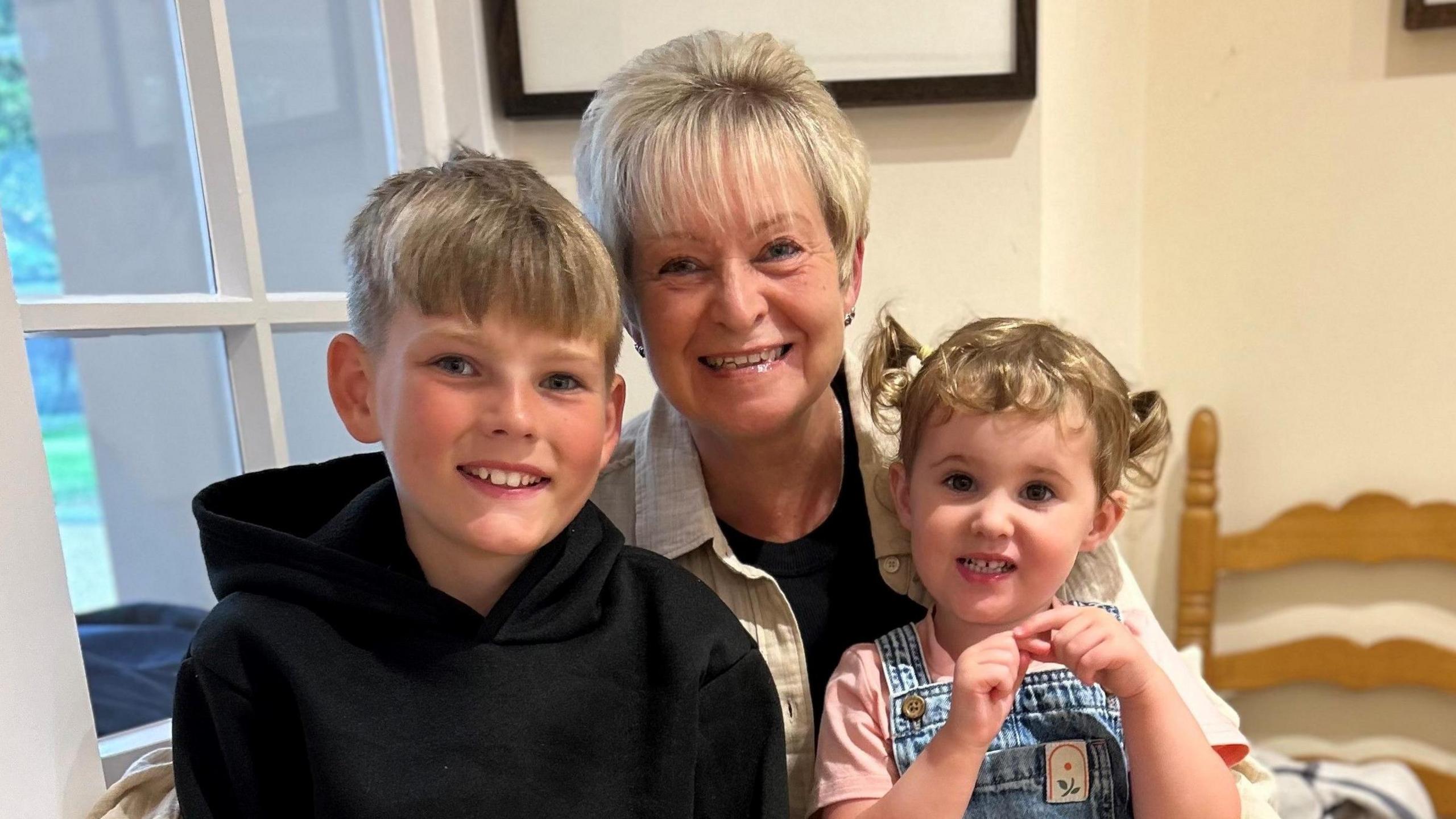 Angela Roberts smiling with her two grandchildren on her lap at a table. Angela has short blonde hair and is wearing a black top with a beige jacket. The children are wearing the same clothes as mentioned above.