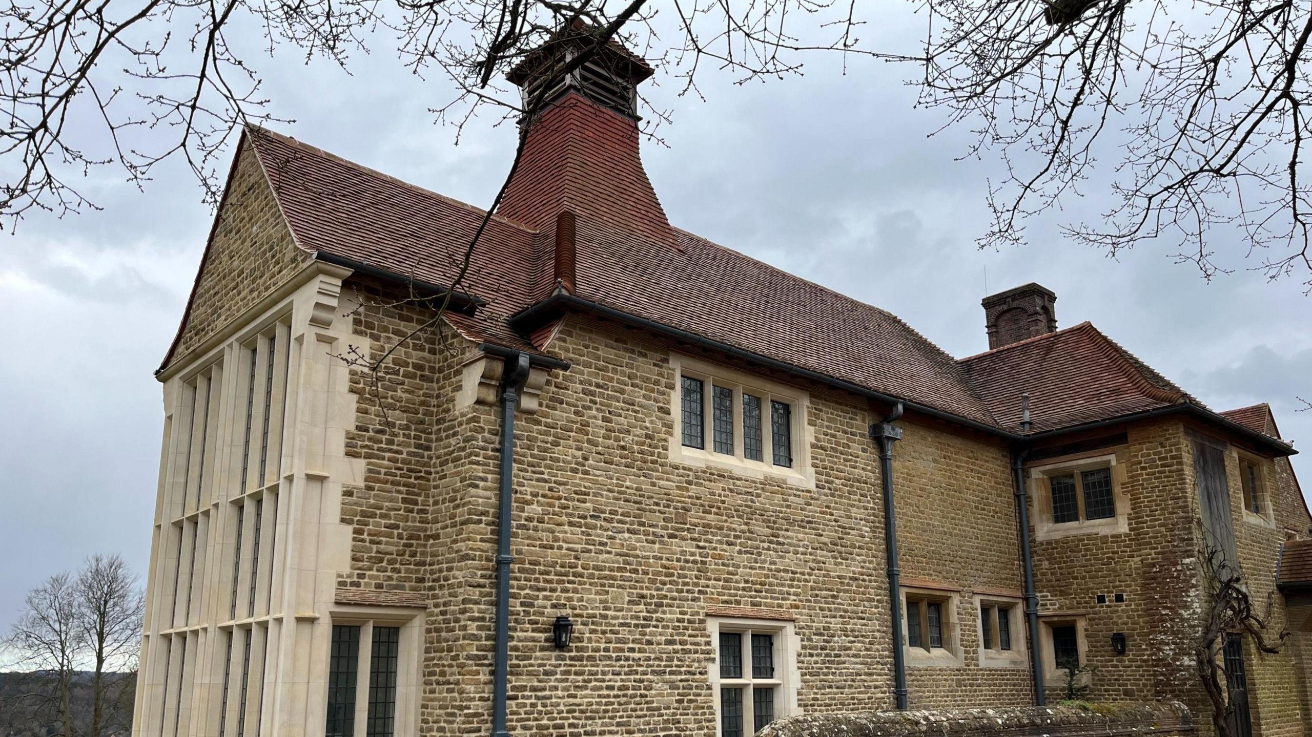 The exterior of a 19th Century country house, with light brown brickwork, a red tile roof and cram stone window fittings.