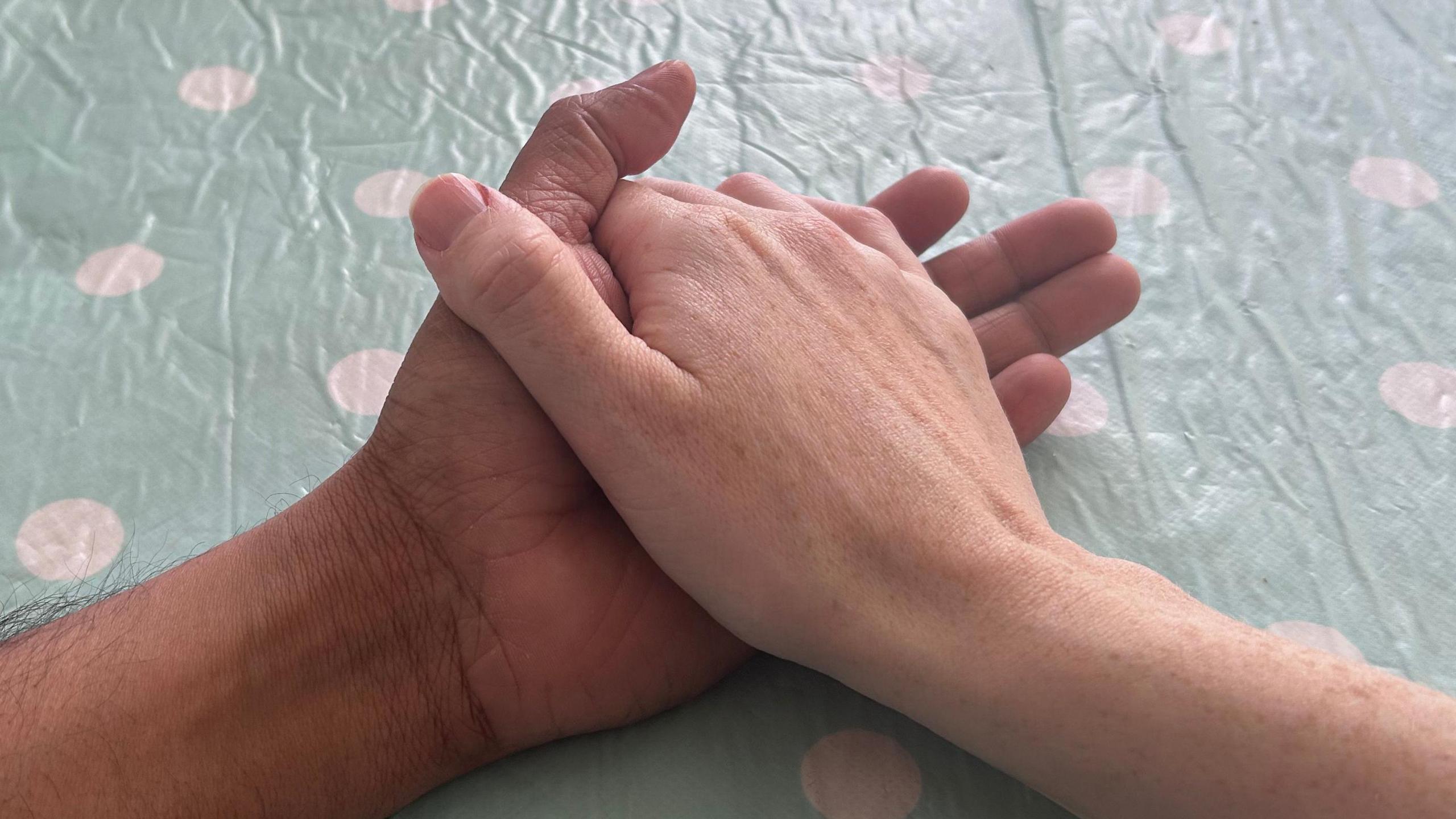 Two arms from two different people resting on a table with a blue and white spotted tablecloth with hands interlinked